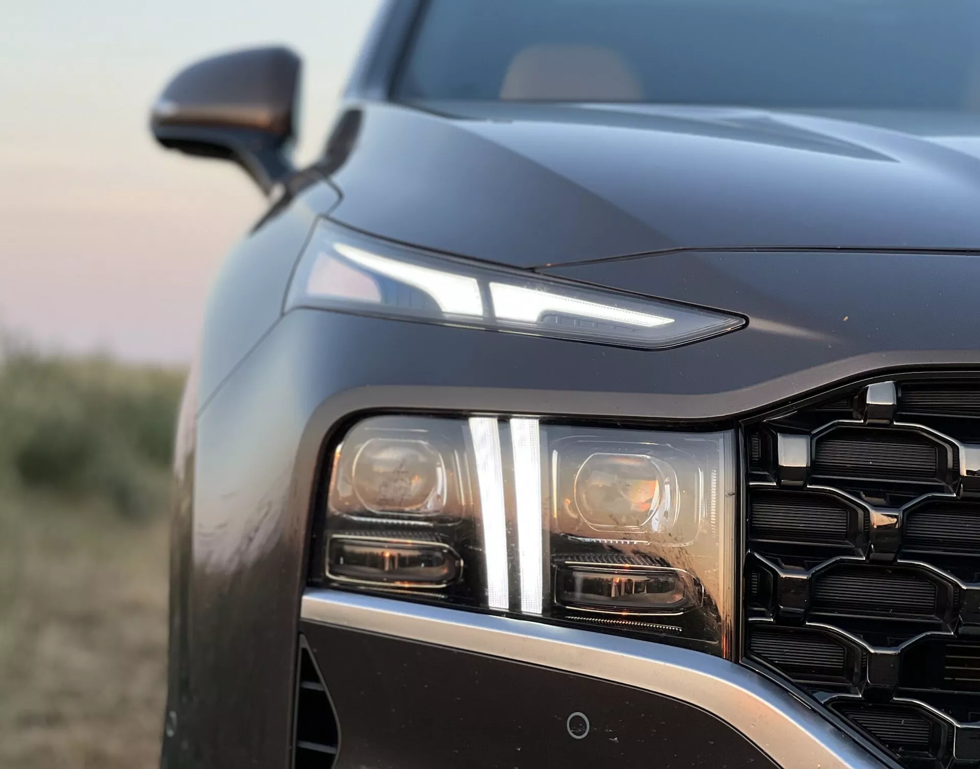 Detailný pohľad na Hyundai Santa Fe 2020, zameriavajúci sa na prednú ľavú časť z mierne predného a nízkeho uhla. Obrázok zdôrazňuje LED alebo HID svetelný klaster s najmenej dvoma samostatnými svetelnými prvkami, pravdepodobne vrátane hlavného lúča a denného svietenia alebo smerovky. Geometricky vzorovaná mriežka a ostro klesajúca línia kapoty naznačujú moderný a agresívny dizajn. Spätné zrkadlo je viditeľné len za líniou kapoty, naznačujúc pokročilé technologické funkcie, ako je senzor alebo kamera v prednom nárazníku, pravdepodobne na asistenciu pri parkovaní alebo vyhnutiu sa kolíziám. Fotografia, urobená počas svitania alebo súmraku, zobrazuje nízkokontrastné osvetlenie s teplými tónmi a plytkú hĺbku ostrosti, ktorá zvýrazňuje dizajnové detaily vozidla na jemne rozmazanom pozadí.