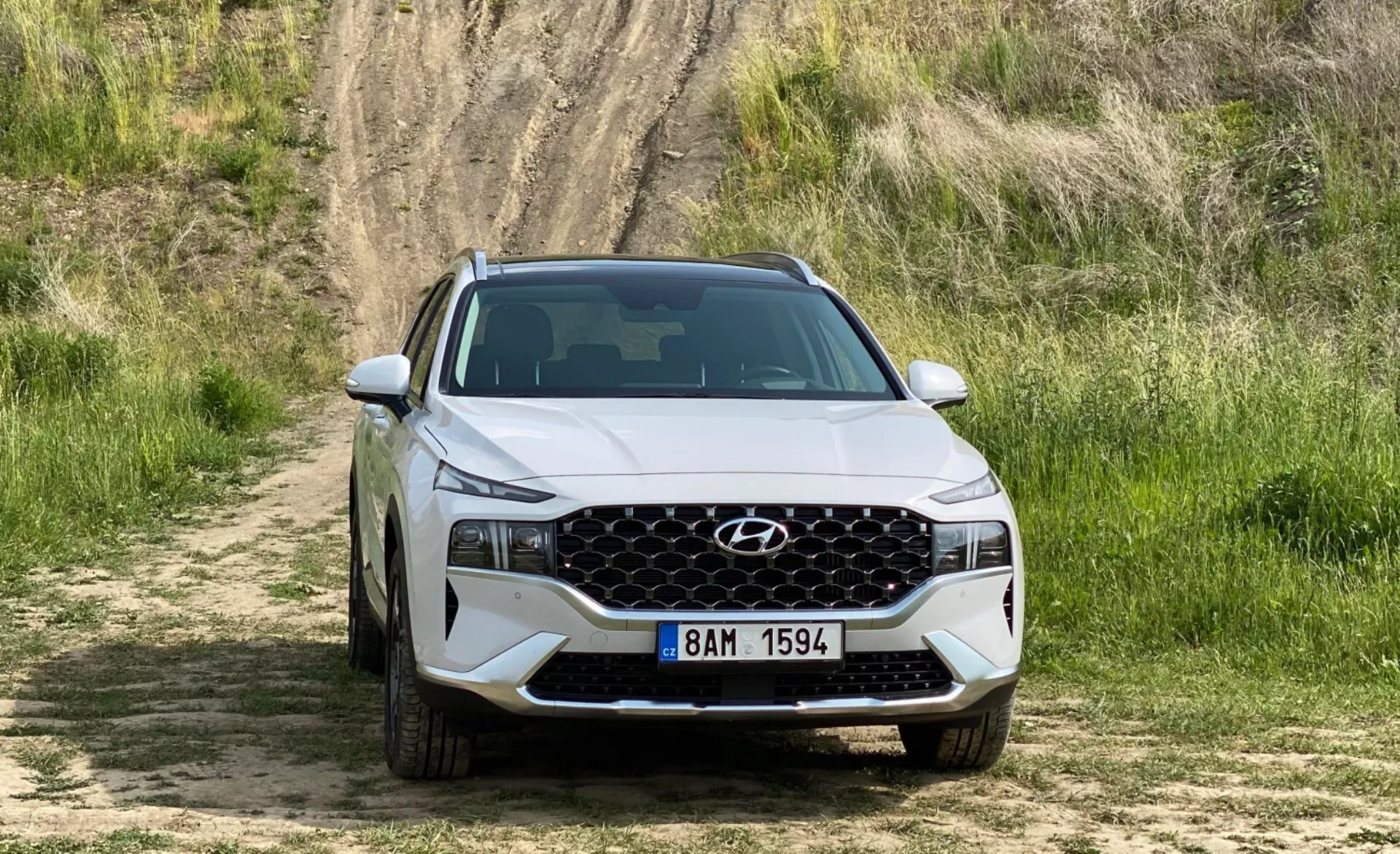 Čelný pohľad na Hyundai Santa-Fe 2020 SUV, ktorý stojí na prašnej ceste s trávou a kopcom v pozadí. Predstavuje prednú mriežku s geometrickým vzorom, ostré moderné svetlomety s LED denným osvetlením a dolný nárazník s otvormi na prívod vzduchu. Kapota má zvýraznené kontúry a je viditeľná európska registračná značka. Obraz zdôrazňuje dizajn a vlastnosti vozidla v terénnom prostredí.