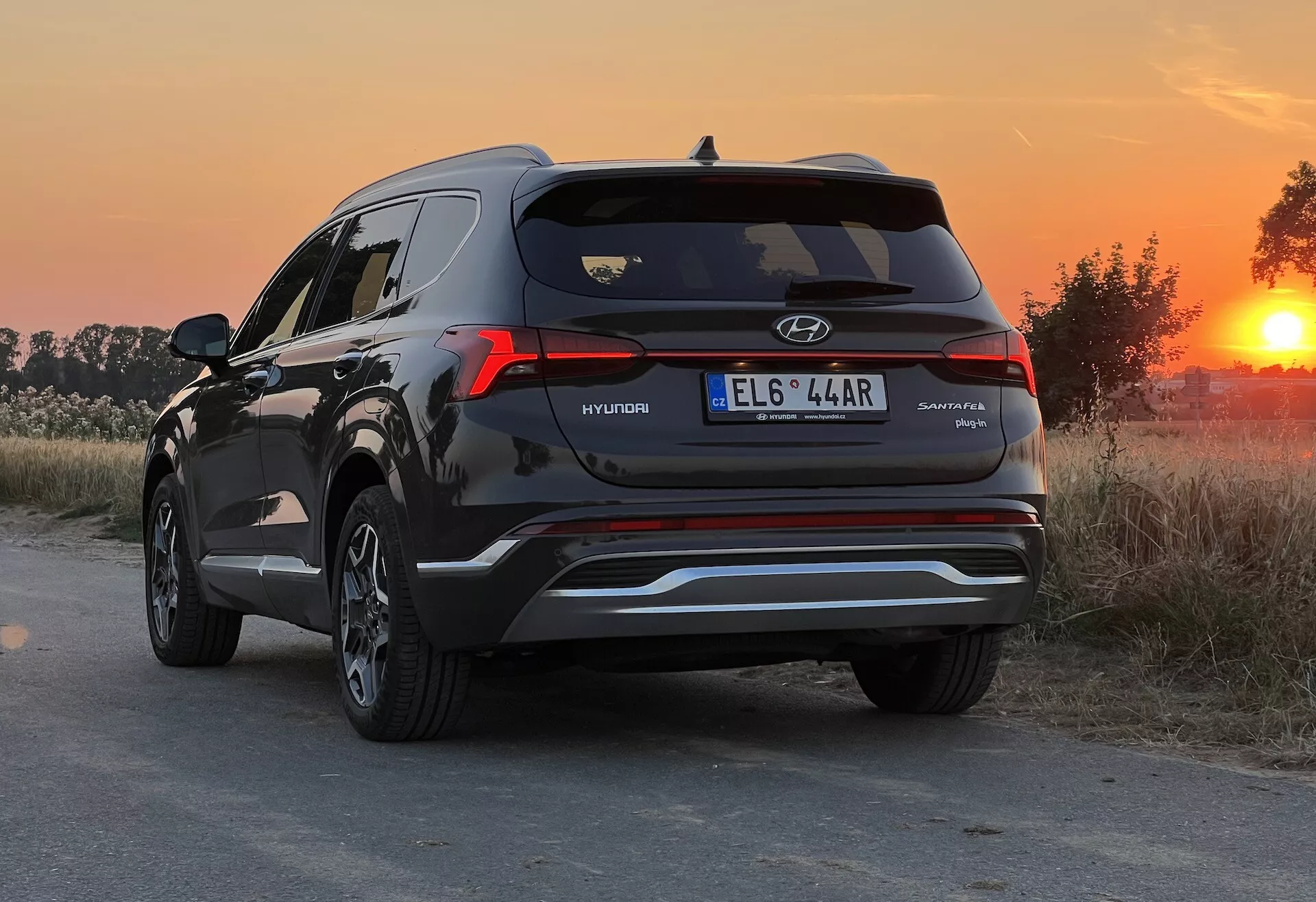 Zadný trojštvrťový pohľad na Hyundai Santa Fe 2020 SUV, zvýrazňujúci pravú stranu a zadný profil s viditeľným označením. Fotografia bola urobená počas zlatej hodiny, ponúkajúc teplé, prirodzené svetlo, ktoré zvýrazňuje farbu laku a odrazy. SUV je zaparkované na prašnej alebo neasfaltovanej ceste v vidieckom prostredí. Nízky uhol kamery, približne vo výške zadných svetiel, zobrazuje dizajn zadných svetiel, zadné okno, kontúry veka kufra a dvojité výfukové koncovky. Viditeľné sú tiež detaily ako kovový akcent na dolnom nárazníku, zadný spojler a žraločia anténa.