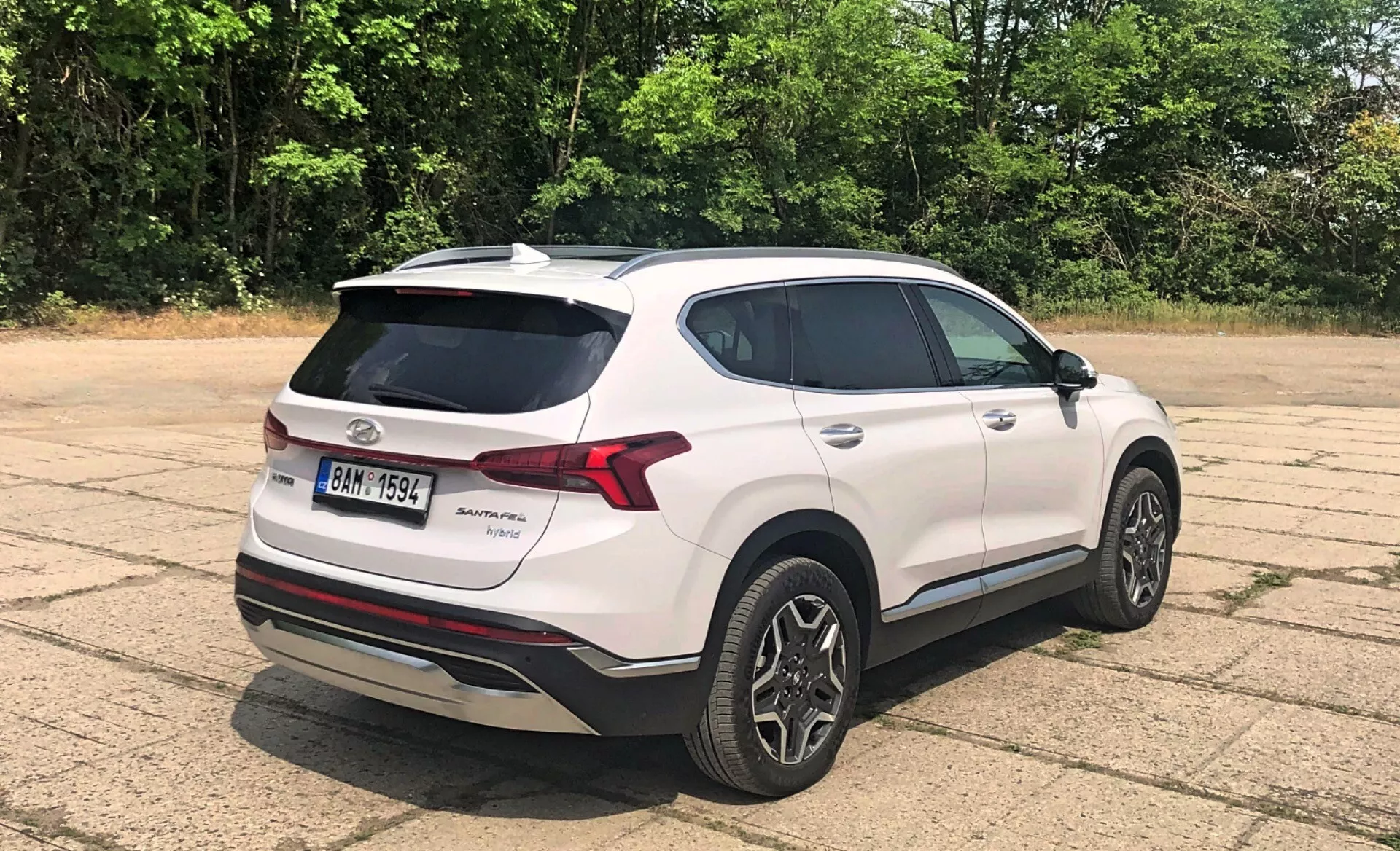 Zadný trojštvrťový pohľad na Hyundai Santa Fe 2020, moderný biely crossover SUV, zaparkovaný vonku na vydláždenej ploche s vegetáciou a stromami v pozadí za slnečného dňa. Viditeľné prvky zahŕňajú LED zadné svetlá, zadný spojler, anténu typu žraločia plutva, dvojfarebné zliatinové kolesá, zatmavené sklá, odznak značky a modelu na zadných dverách, strieborný ochranný plech pod zadným nárazníkom, bočné zrkadlá vo farbe karosérie s integrovanými smerovými svetlami a chrómované lišty okolo okien.