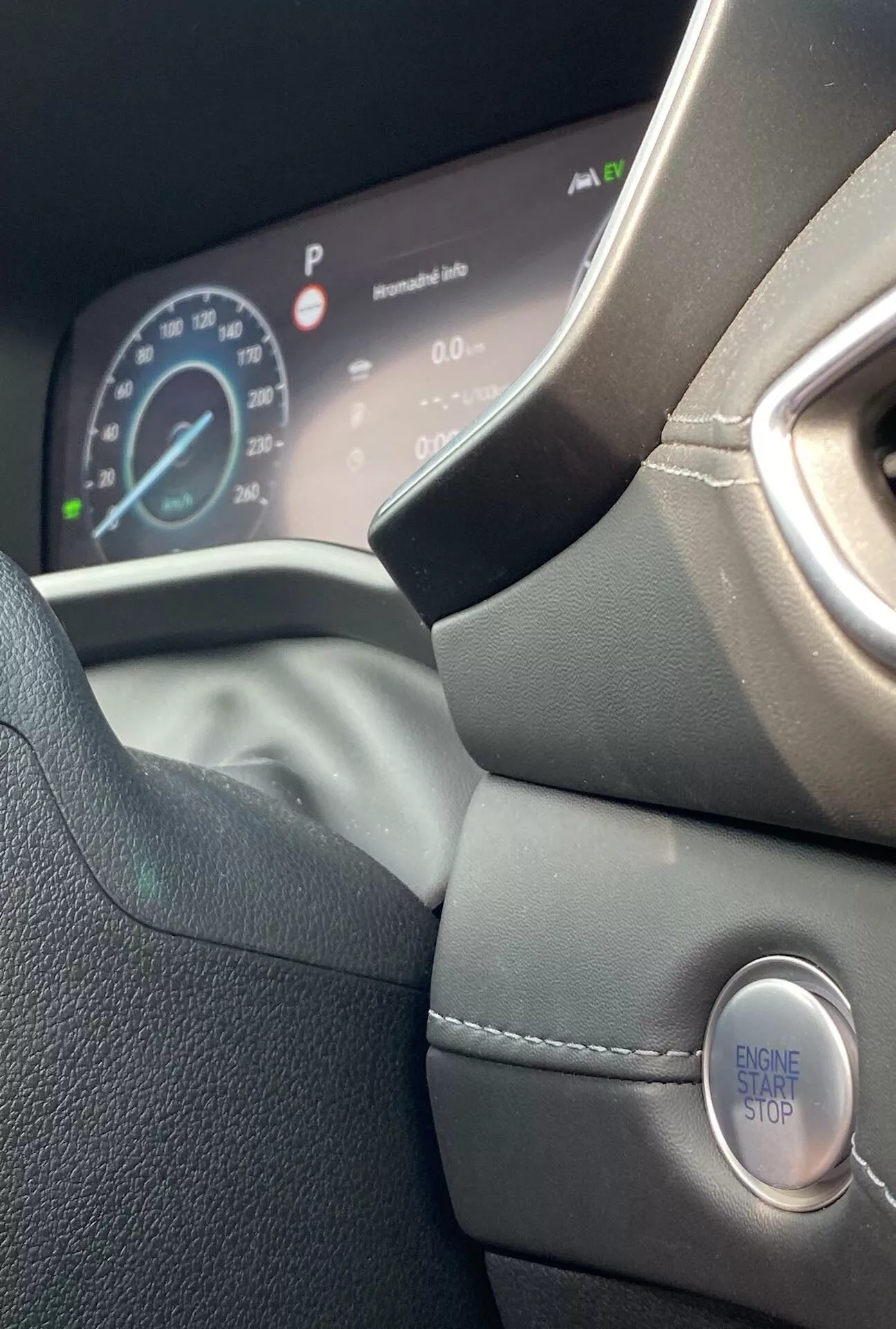 Detailný pohľad na interiér modelu Hyundai Santa Fe 2020, zameraný na ľavú stranu koženého volantu a digitálnu prístrojovú dosku. Fotka urobená z miesta vodiča a mierne naklonená nahor ukazuje moderný dizajn volantu, detaily prešívania, tvarovaný úchyt a bezkľúčové zapaľovanie s tlačidlom 'ENGINE START STOP'. Digitálna prístrojová doska zobrazuje rýchlomer vozidla, ktorý ukazuje, že auto stojí, a ďalšie informácie o vozidle, zdôrazňujúc prémiový a ergonomický dizajn.