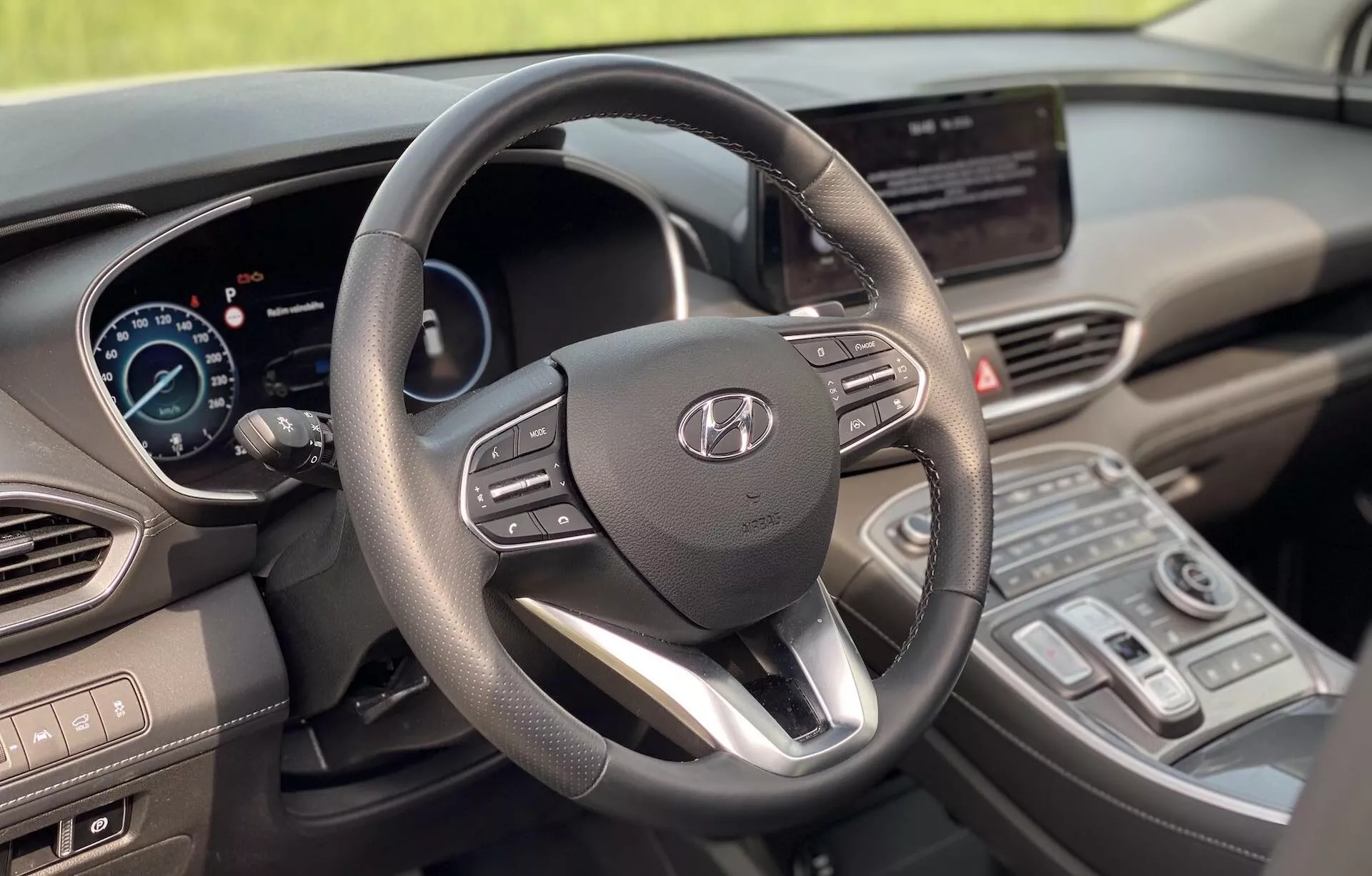 Interiér Hyundai Santa Fe 2020 z pohľadu vodiča, zameraný na moderný trojramenný volant s logom Hyundai a integrovanými ovládačmi, digitálny prístrojový panel, centrálny dotykový infotainment displej, centrálnu konzolu s ovládačmi klimatizácie a modernú radiacu páku. Dizajn ukazuje rovnováhu medzi funkčnosťou a modernou estetikou s mäkkými materiálmi a kovovými akcentmi, čo naznačuje pokročilé technologické vybavenie. Obrázok zobrazuje čisté, dobre udržiavané, stacionárne vozidlo bez rozmazania.