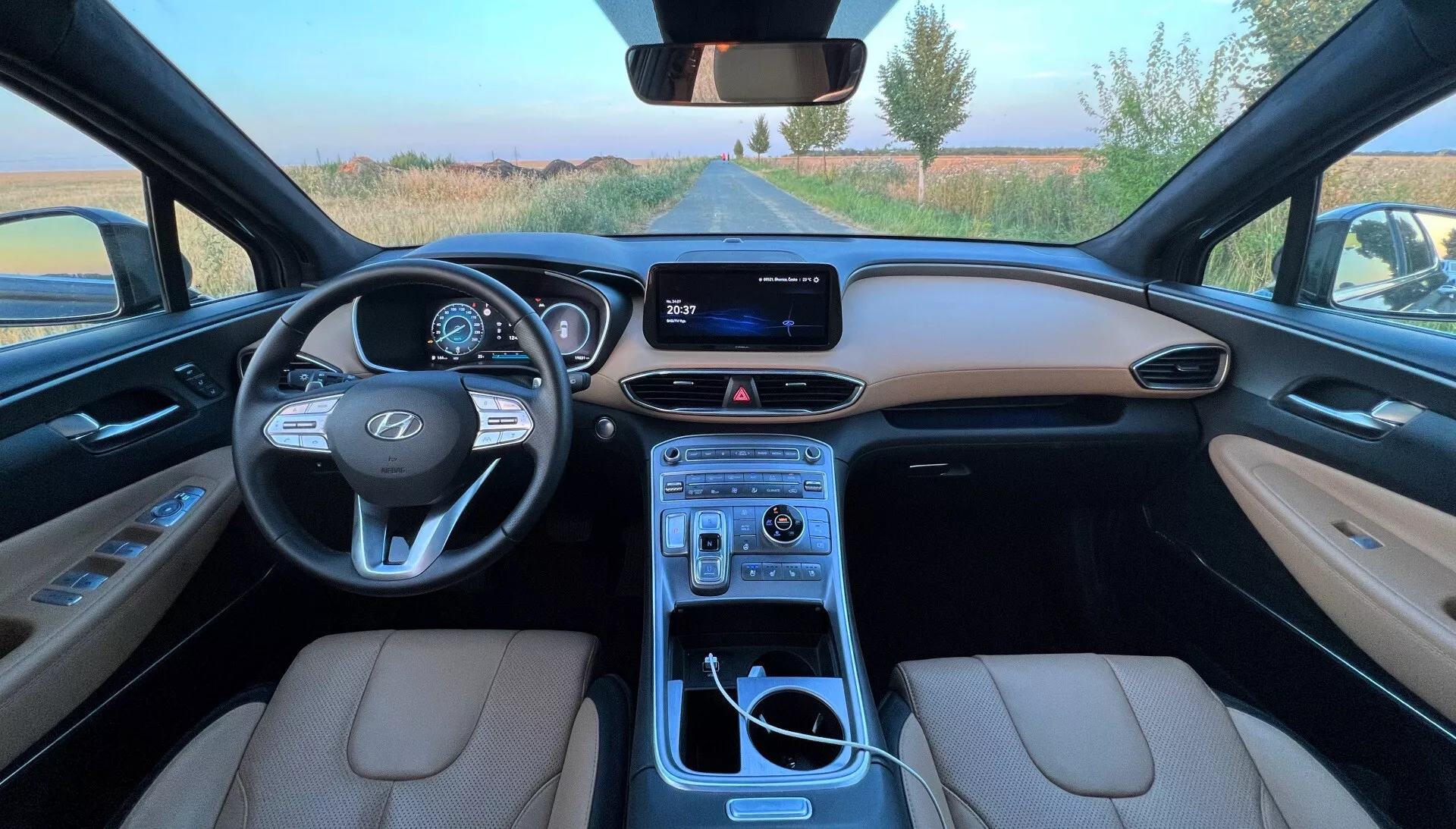 Širokouhlý pohľad z vodičovho sedadla na Hyundai Santa Fe 2020, ukazujúci béžové kožené sedadlá, detailný prístrojový panel s digitálnym displejom, volant Hyundai s ovládacími prvkami, infotainment systém s displejom a plne vybavenú stredovú konzolu s radiacou pákou, držiakmi na poháre, ovládacími gombíkmi a úložným priestorom.