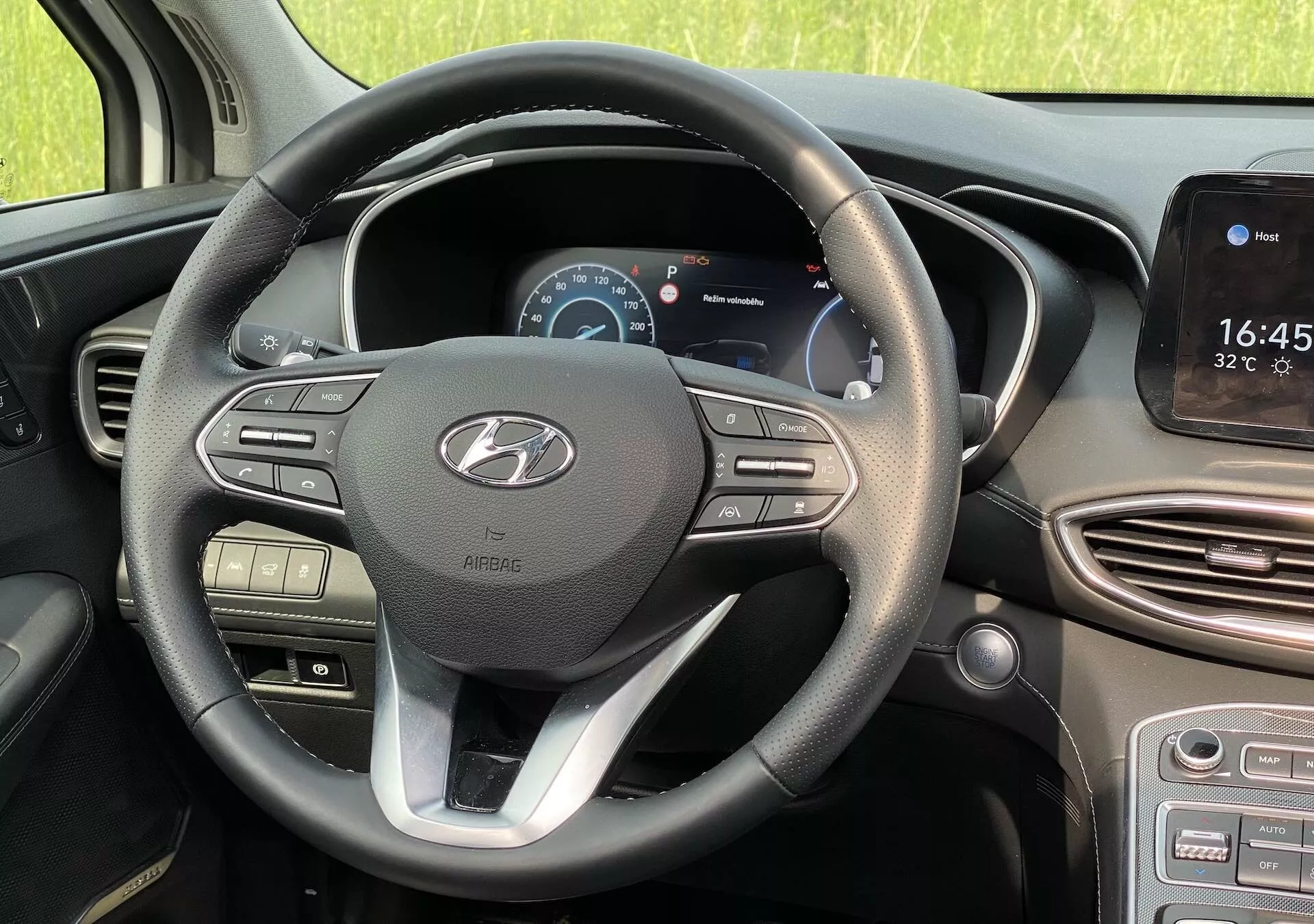 Pohľad na interiér vozidla Hyundai Santa Fe 2020 z pohľadu vodiča, ukazujúci moderný trojramenný volant so strieborným akcentom a logom Hyundai. Na ľavom ramene sa nachádza ovládanie hlasitosti a médií, zatiaľ čo pravé rameno má tlačidlá pre tempomat a navigáciu v menu. Za volantom sa nachádza digitálny prístrojový panel zobrazujúci rýchlosť a otáčkomer. Vpravo je dotyková obrazovka infotainmentu zobrazujúca čas a teplotu, s možnou navigáciou a integráciou smartfónu. Pod ňou sú vetracie otvory a tlačidlá klimatizácie. Vozidlo je zaparkované za denného svetla s prírodným, neurbánnym prostredím viditeľným cez okná.