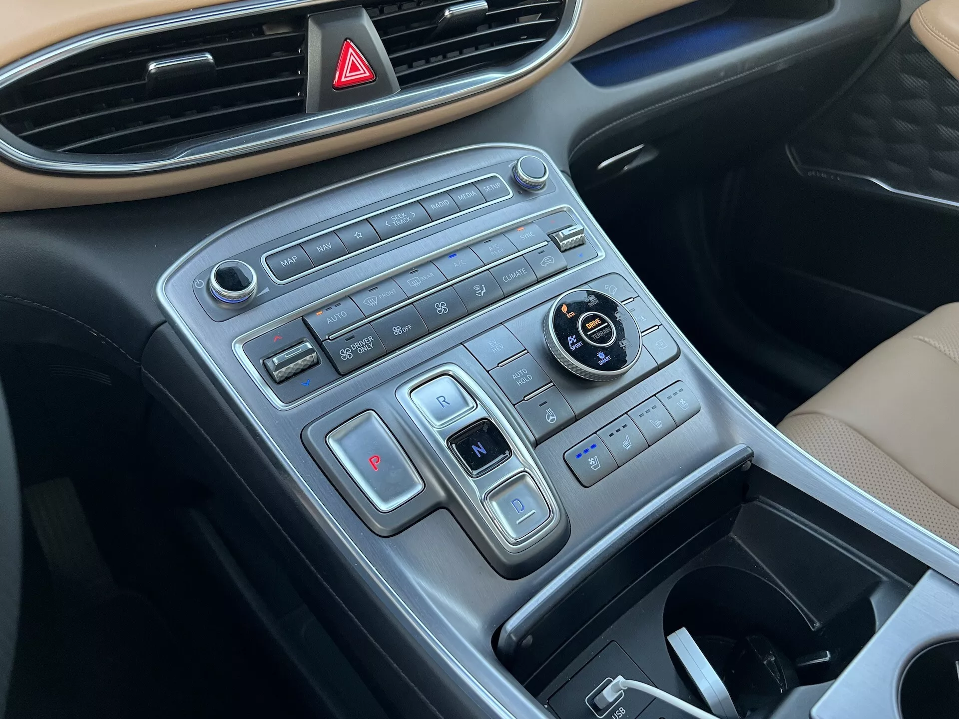 Detailný pohľad na stredovú konzolu Hyundai Santa Fe 2020 z pohľadu pasažiera, ktorý zobrazuje prístrojovú dosku s tlačidlom výstražných svetiel v hornej časti, moderný dizajn ovládačov v horizontálnych radoch vrátane voliča režimu jazdy, nastavovania trakčnej kontroly, dvojzónového klimatizačného systému, tlačidiel vyhrievania sedadiel a zásuvnú úložnú priehradku, s povrchovou úpravou pripomínajúcou brúsený kov.