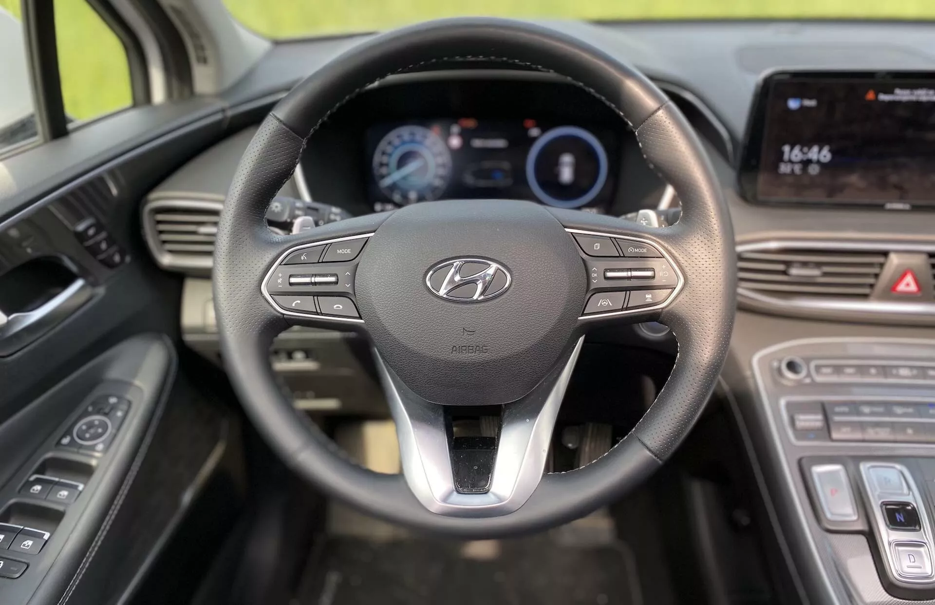 Pohľad na interiér vozidla Hyundai Santa-Fe 2020 z pohľadu vodiča, kde je vidieť výrazný volant s logom Hyundai a ovládacími tlačidlami pre multimédiá a konektivitu. Za volantom sa nachádza osvetlený digitálny prístrojový panel zobrazujúci kľúčové informácie ako rýchlosť a spotreba paliva. Vpravo je stredová konzola s dotykovým displejom, ktorý ukazuje čas (16:46) a pravdepodobne podporuje funkcie navigácie a multimédií. Pod displejom sú vidieť dotykové ovládanie klimatizácie, čo zvyšuje moderný dizajn vozidla. Na ľavej strane volantu sú vidieť ovládacie prvky dverí pre elektrické okná a nastavenie bočných zrkadiel.