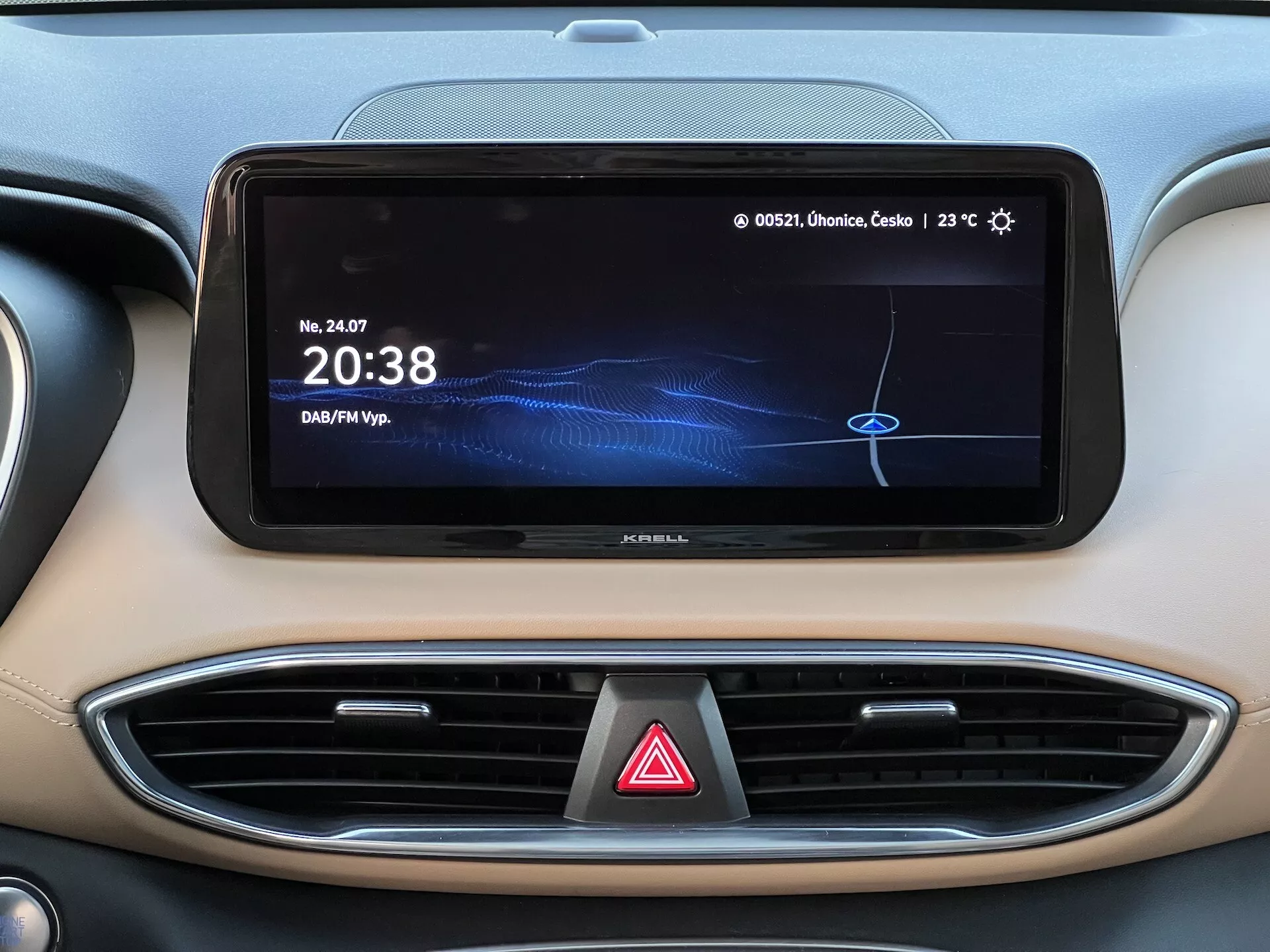 Detailný záber na stredovú časť palubnej dosky v Hyundai Santa Fe 2020, zobrazujúci široký horizontálny dotykový displej infotainmentu s digitálnymi hodinami '20:38', zdroj média označený 'DAF/MP3', údaj o teplote '23°C' a čiastočne viditeľným poľským textom. Viditeľné sú aj dva ventilačné otvory s chrómovým prízvukom a tlačidlo výstražných svetiel pod displejom. Palubná doska má dvojfarebný farebný vzor s vysokokvalitným moderným vnútorným dizajnom.