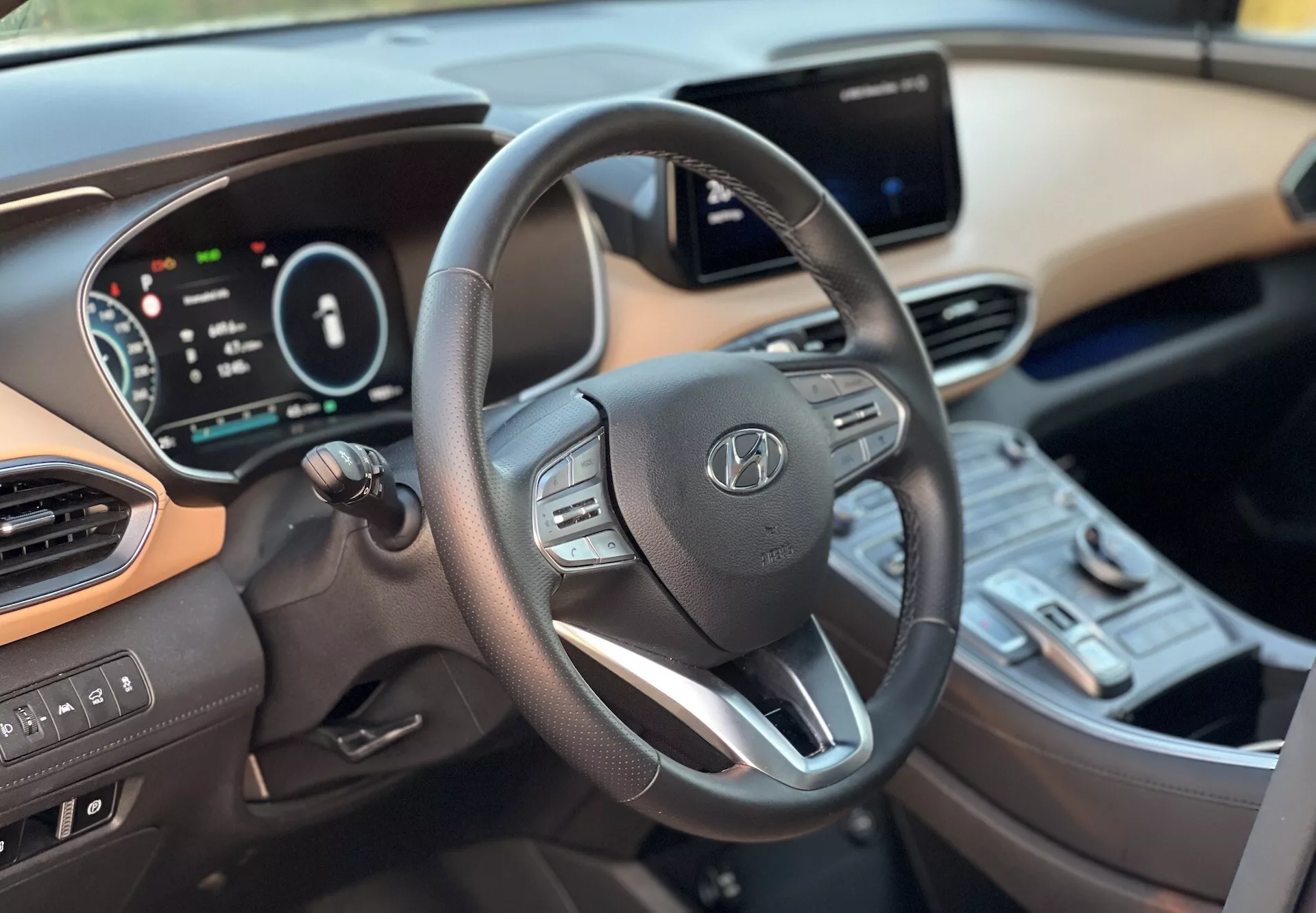 Interiérový pohľad na Hyundai Santa-Fe 2020 zobrazujúci vodičovú stranu z pravého stredu, zameraný na moderný trojramenný volant s ovládacími prvkami, digitálny prístrojový panel a dotykový infotainment systém v centre konzoly. Viditeľné sú aj spínače klimatizácie, tlačidlo štartovania a dvojfarebné schéma prístrojovej dosky a interiéru, ilustrujúce pokročilú technológiu a pohodlie vozidla.