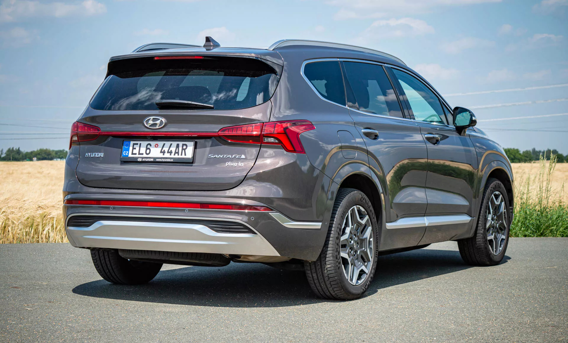 Trojštvrťový zadný pohľad na Hyundai Santa Fe Hybrid 2020. Záber z nízkeho uhla zdôrazňuje výšku a robustnosť SUV, vozidlo je umiestnené vpravo, čo naznačuje pohyb. Zaparkované na vydláždenej ceste v vidieckom prostredí, zadná časť má vertikálne usporiadané LED zadné svetlá, zadný spojler pre aerodynamickú stabilitu, dvojité výfukové koncovky, delený strešný spojler a reflexné lišty na nárazníku. Dizajn naznačuje zadné parkovacie senzory, zadnú kameru, viaclúčové zliatinové kolesá a žraločiu plutvovú anténu pre satelitné rádio alebo GPS technológiu.