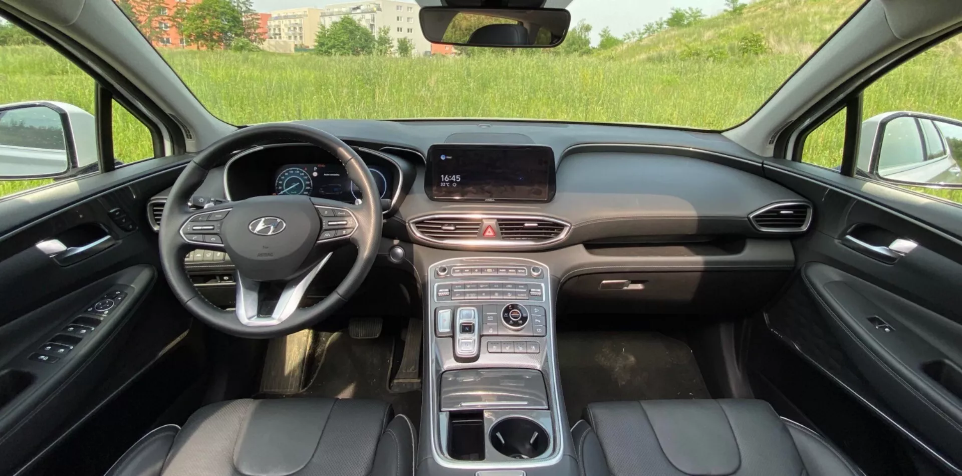 Pohľad na interiér Hyundai Santa Fe 2020, ukazujúci modernú prednú kabínu s centrálnou palubnou doskou vybavenou digitálnym informačným displejom, elegantným vetracím pásom a multifunkčnými tlačidlami na volante. Dizajn volantu pre jazdu vľavo zahŕňa výrazné logo výrobcu a pravdepodobne digitálny prístrojový panel. Stredová konzola má otočný volič prevodov, držiaky na nápoje a pravdepodobne aj opierku na ruky. Predné sedadlá sú čalúnené čiernou kožou s viditeľným prešívaním, čo naznačuje elektrické nastavovanie a luxus. Prostredie sa zdá byť vonku za denného svetla, s mierne rozmazaným zeleným poľom viditeľným cez čelné sklo.