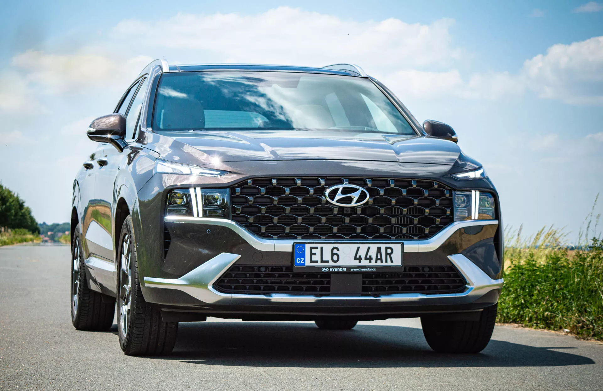 Predný uhlový pohľad na moderné SUV, konkrétne Hyundai Santa Fe 2020, urobený z nízkeho pohľadu blízko strany vodiča. Kamera zachytáva odvážny predný dizajn vozidla, vrátane metalickej čiernej alebo tmavošedej farby s lesklým leskom, mriežky s medovou štruktúrou a centrálnym emblémom Hyundai, ostrých uhlových svetlometov s LED dennými svetlami, prídavné dolné osvetlenie ako hmlové svetlá, veľkého nasávacieho otvoru a tvarovaných línií kapoty. Predné sklo je veľké a sklonené, s čiastočne viditeľnými bočnými zrkadlami s integrovanými smerovými svetlami. ŠPZ je namontovaná v strede dolnej časti nárazníka.