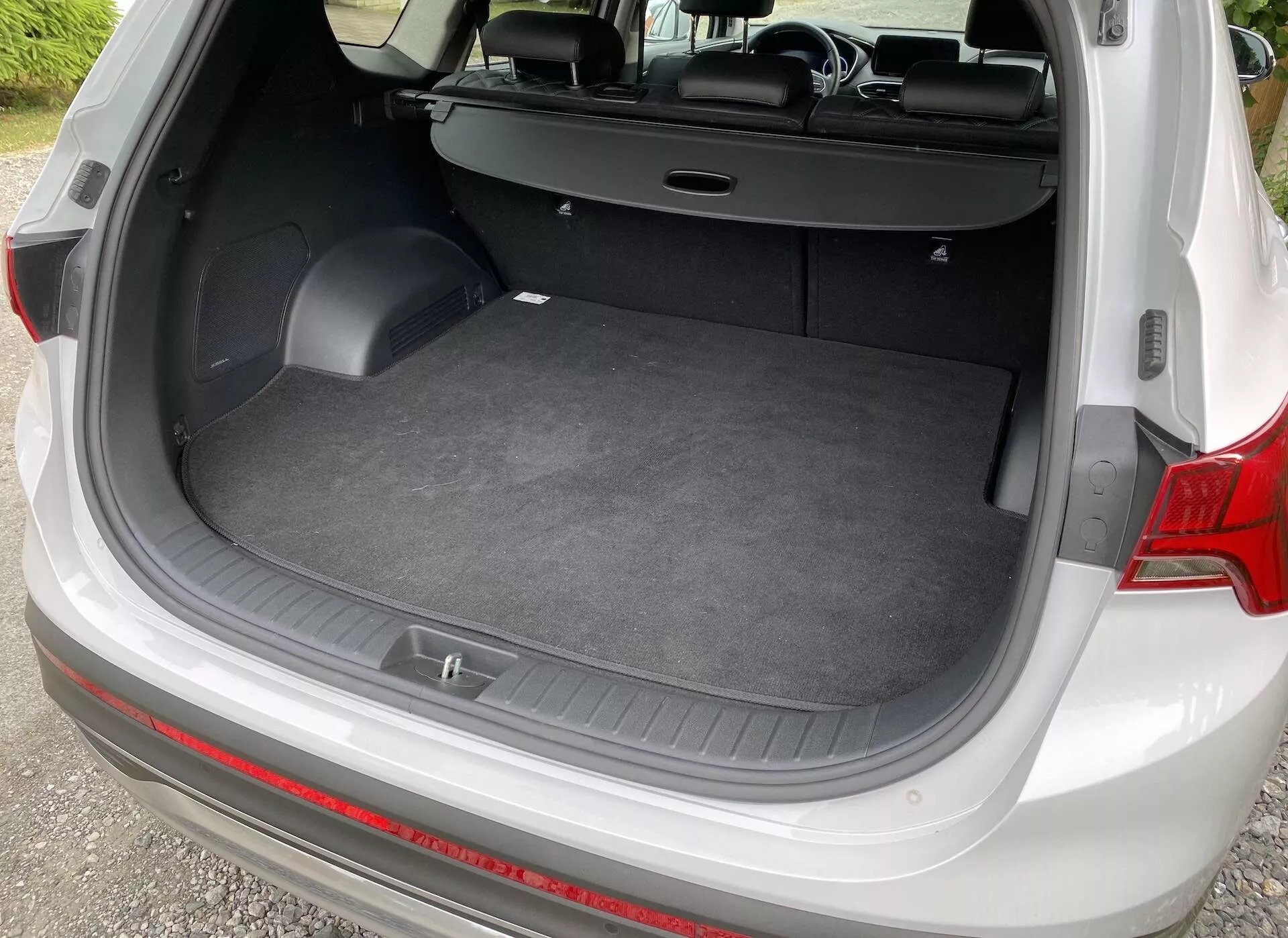 Pohľad z vysokého uhla na batožinový priestor Hyundai Santa-Fe 2020, zobrazený zozadu s otvoreným poklopom. Obrázok ukazuje rovný nakladací priestor, koberčekovú podlahu a bočné steny obsahujúce malé úložné priehradky a háčiky na príslušenstvo. Zadné sedadlá sú sklopné do vzpriamenej polohy a výsuvný kryt batožinového priestoru je zasunutý dozadu, čo zdôrazňuje všestranný interiér vozidla.