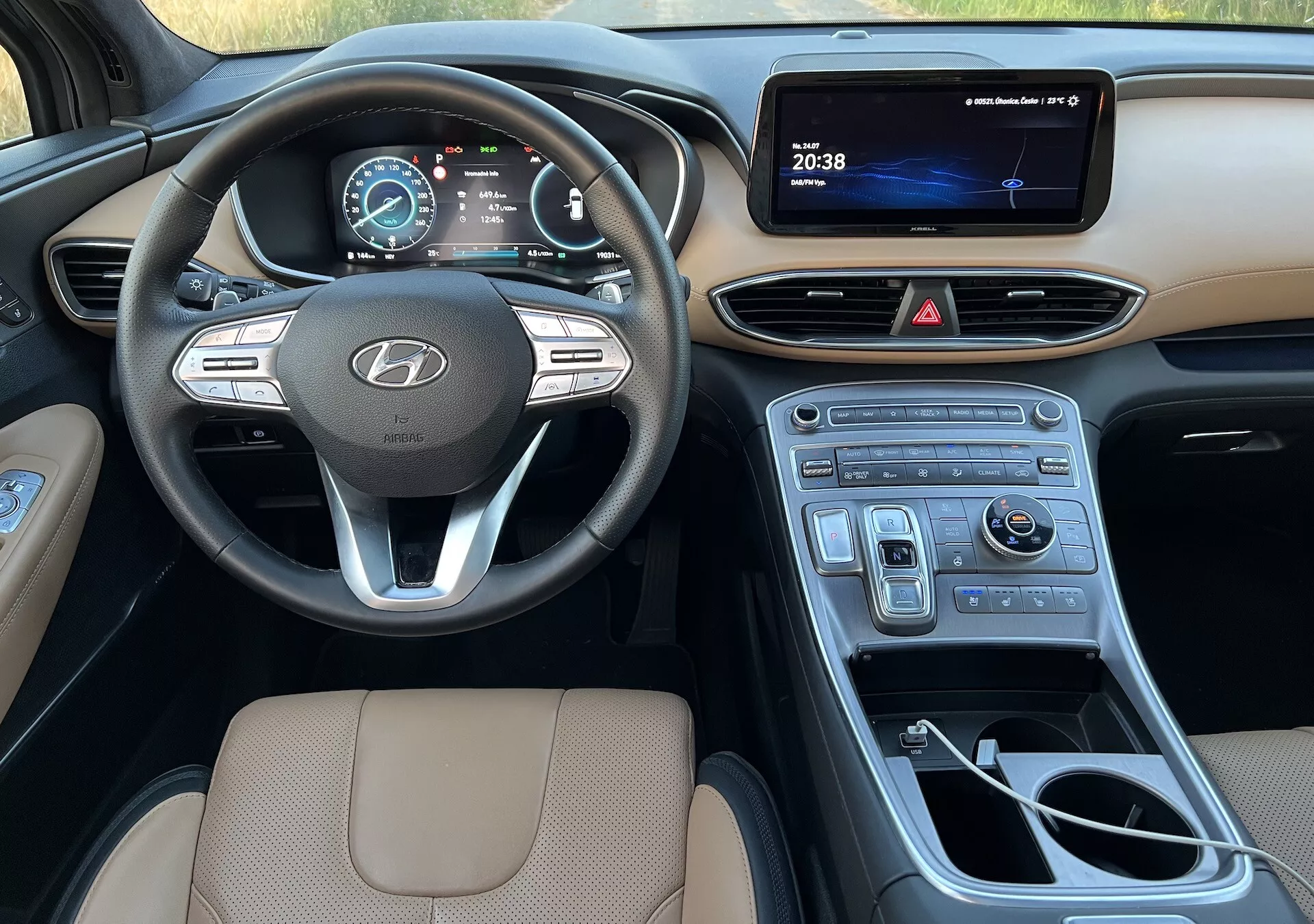 Tento obrázok ukazuje interiér Hyundai Santa-Fe 2020, zameriava sa na stranu vodiča. Poskytuje komplexný pohľad od sedadla vodiča po stredovú konzolu a rozširuje sa na stranu pasažiera, zdôrazňujúc niekoľko kľúčových prvkov. Volant s logom Hyundai, s namontovanými ovládacími prvkami pre audio, tempomat a systém hands-free, je výrazne zobrazený. Za ním je digitálny prístrojový panel zobrazujúci tachometer a informácie o vozidle. Stredová konzola zahŕňa displej infotainmentu, ovládacie tlačidlá pre výstražné svetlá a klimatizáciu, a lesklý drevený obklad. Tiež je vidieť radiacu páku pre automatickú prevodovku, ovládací gombík pre navigáciu alebo multimédiá a držiaky na nápoje. Horná časť sedadla vodiča, čalúnená v béžovej farbe s prešitím, zdôrazňuje pohodlie a kvalitu dizajnu. Obrázok zachytáva moderný, užívateľsky prívetivý dizajn a technologické prvky interiéru vozidla počas denného svetla s prirodzeným osvetlením.