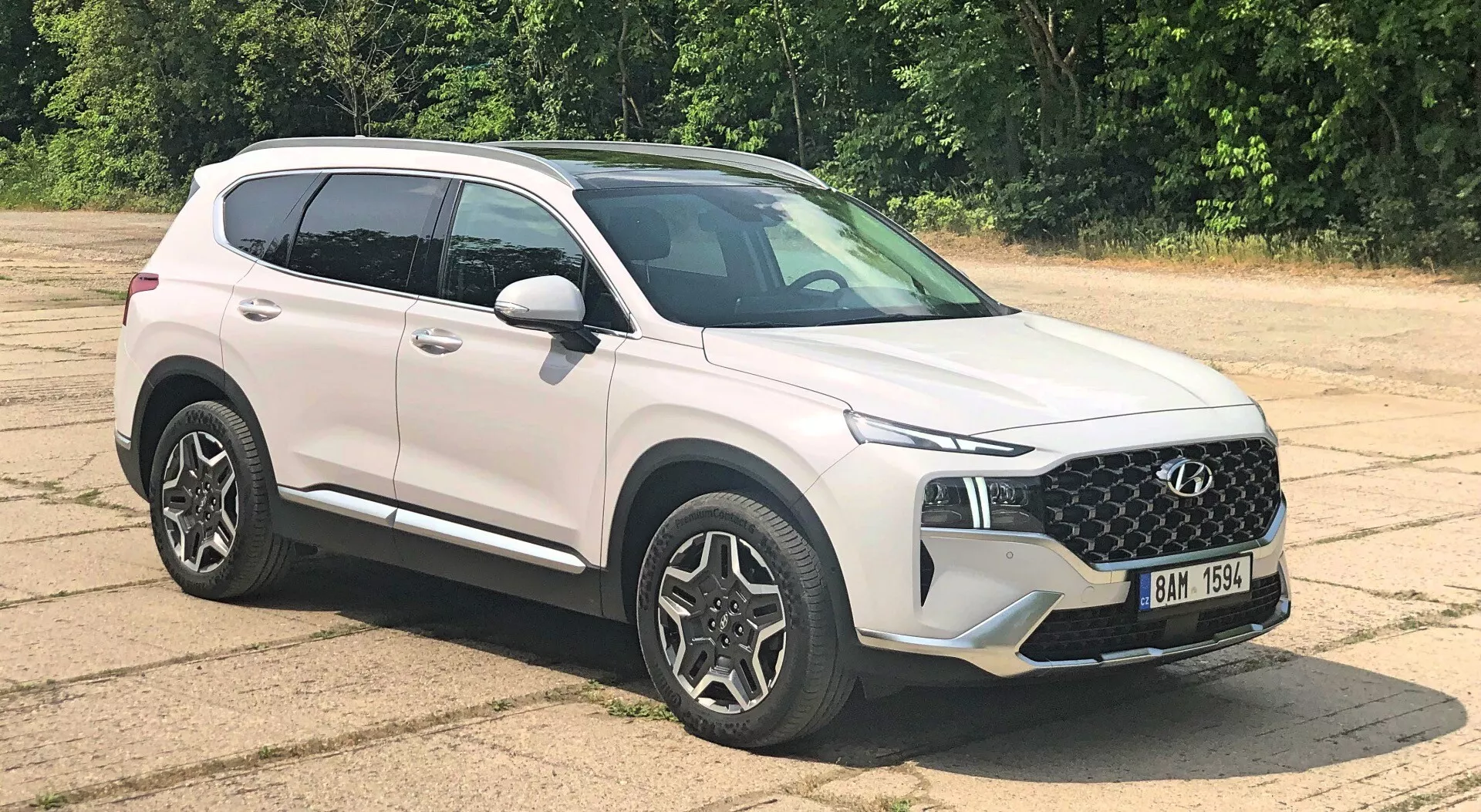 Moderné SUV Hyundai Santa Fe 2020 zaparkované na asfaltovom povrchu z pohľadu na tri štvrtiny, zobrazujúce dizajn masky, elegantné svetlomety, viaclúčové zliatinové kolesá, spätné zrkadlá vo farbe karosérie so zabudovanými smerovkami, charakterové línie, aerodynamický profil, lesklý biely lak, mierne zatemnené okná a prirodzené, rovnomerné osvetlenie s vegetáciou v pozadí.