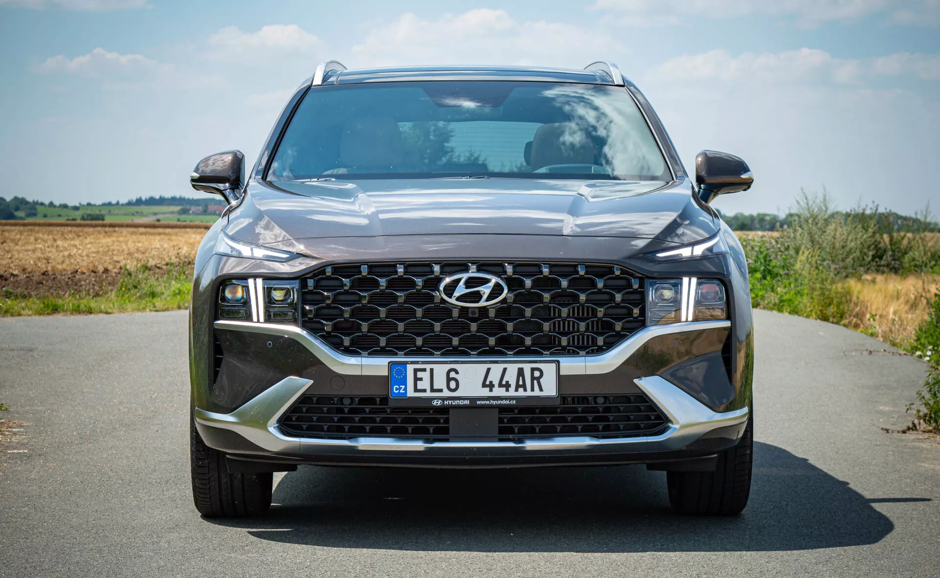 Hyundai Santa Fe 2020 - Čelný pohľad na moderné SUV na asfaltovej ceste s prírodnou krajinou v pozadí. Tento mierne nízky uhol zvýrazňuje impozantný vzhľad SUV, zdôrazňujúc detaily ako šesťuholníkovú prednú mriežku so vzorom voštiny, asymetrické LED svetlá, kontrastné prvky predného nárazníka a ďalšie osvetľovacie prvky, vrátane hmlových svetiel.