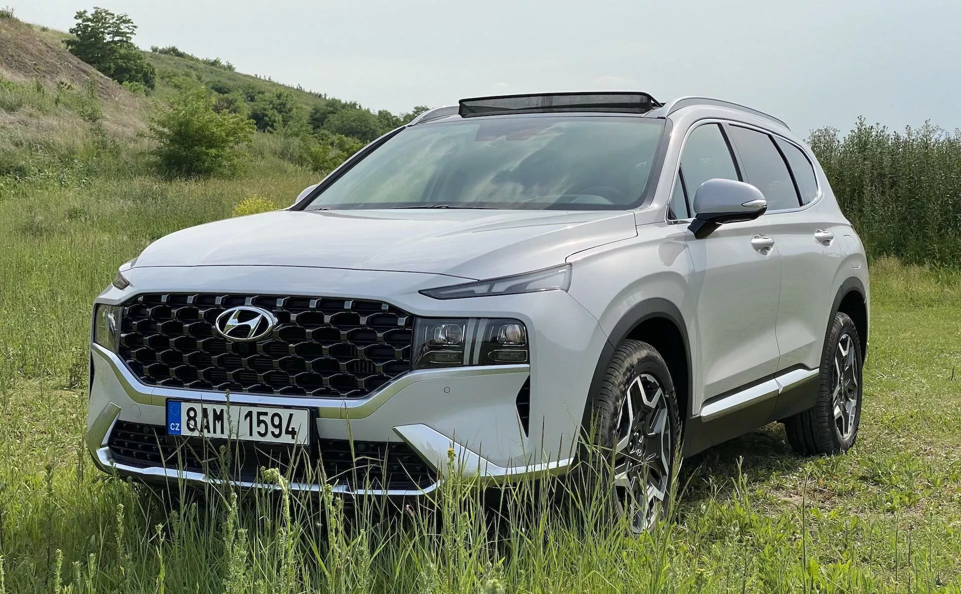Moderné SUV Hyundai Santa Fe 2020 v prírodnom, vonkajšom prostredí. Vozidlo je zaparkované na trávnatej ploche s ľahkým sklonom, čo naznačuje terénne schopnosti. Svetlý denný záber ukazuje predný a bočný profil s črtami ako výrazná geometrická mriežka, úzke LED svetlomety, denné svetlá, zliatinové kolesá s viac paprskovým dizajnom, bočné zrkadlá v farbe karosérie s integrovanými smerovkami, strešné lišty a plastové clony okolo blatníkov. Záber zachytáva dizajn a dobrodružný potenciál SUV, zvýrazňujúc jeho užitočnosť a prémiový vzhľad.