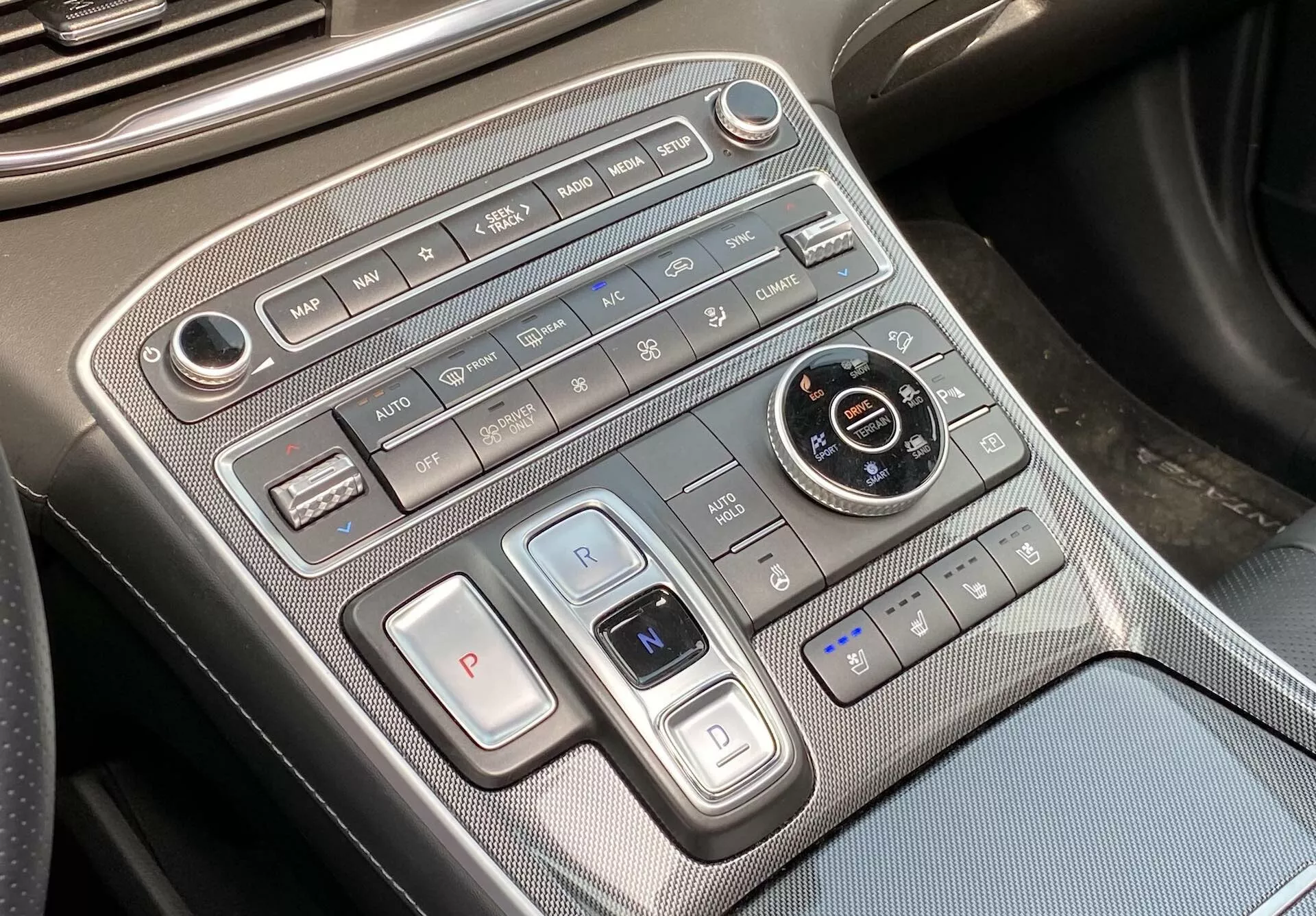 Tento obrázok ukazuje detailný pohľad na stredovú konzolu Hyundai Santa Fe 2020, zameriavájúci sa na ovládanie klímy a prevodovky. Záber, ľahko uholovaný z pohľadu vodiča alebo spolujazdca, odhaľuje horizontálne usporiadaný panel ovládania klímy na vrchu, obsahujúci ovládanie rýchlosti ventilátora, tlačidlá pre smer prúdenia vzduchu, automatickú klimatizáciu, odmrazovanie a recirkuláciu vzduchu. Nižšie sa nachádzajú tlačidlá na výber prevodovky s veľkým tlačidlom 'P' pre parkovanie a prepínačom na prevodové stupne 'R' a 'N', naznačujúcim použitie tlačidlového alebo elektronického systému pre výber prevodovky. K dispozícii je tiež okrúhle koliesko na výber jazdných režimov, obklopené tlačidlami pre automaické držanie brzd, parkovacieho asistenta a systému štart/stop. Kontroly pre vyhrievané alebo odvetrávané sedadlá sú viditeľné v pravom dolnom rohu. Konzola má prémiové materiály s kovovými akcentmi a vzorom podobným uhlíkovým vláknam. USB port v ľavom dolnom rohu ponúka možnosti pripojenia zariadení.