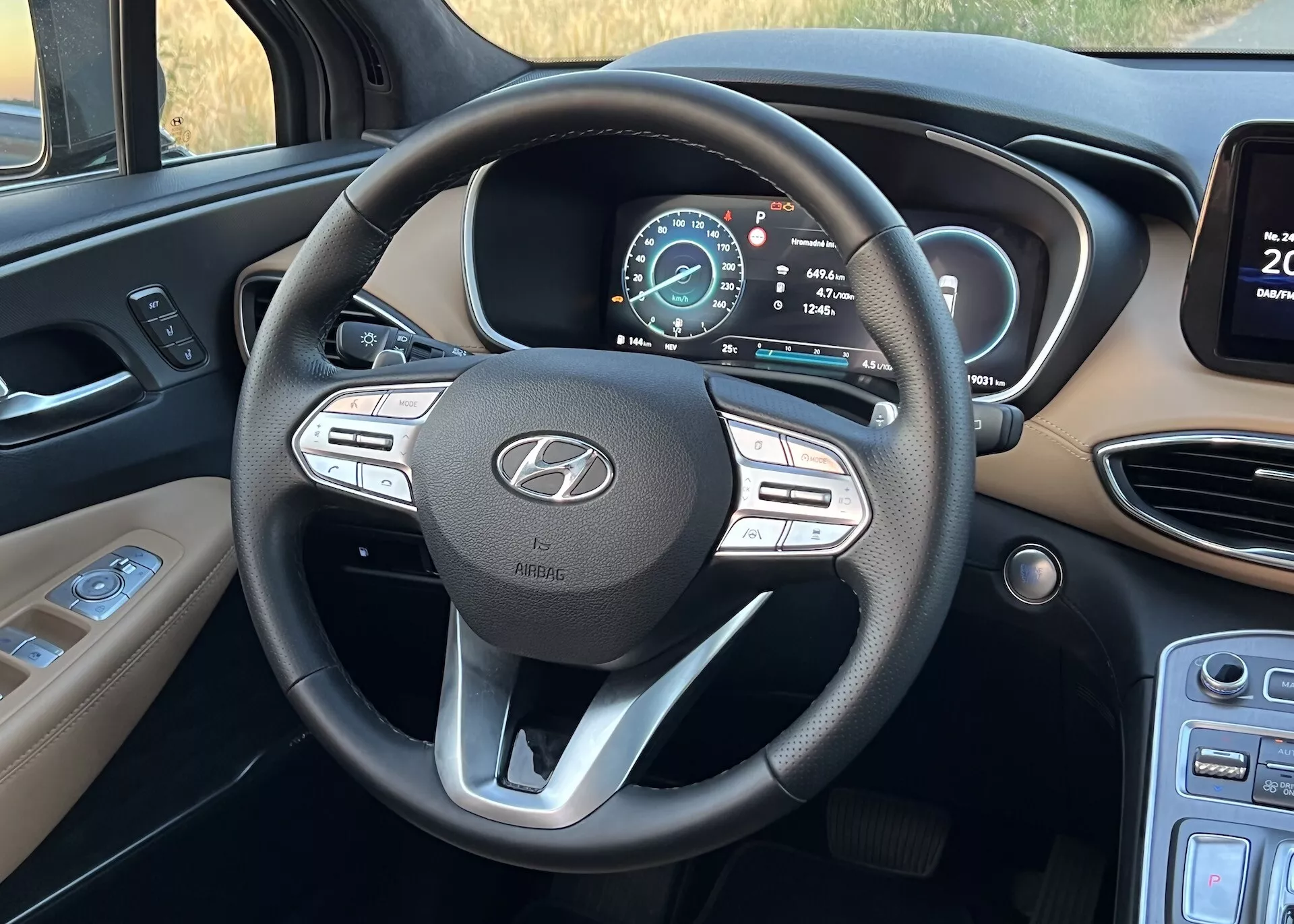 Fotografia interiéru Hyundai Santa Fe 2020 zameraná na multifunkčný trojramenný volant a digitálny prístrojový panel. Fotografia je zhotovená z pohľadu sedadla vodiča a zobrazuje ovládacie prvky médií a tempomatu na volante, odznak Hyundai s textom 'AIRBAG' a multifunkčný digitálny prístrojový panel, ktorý naznačuje, že vozidlo je v parkovacom režime a dvere sú pootvorené. Naľavo sú pravdepodobne ovládacie prvky svetiel, napravo je viditeľný okraj informačného systému spolu s vetracími otvormi.
