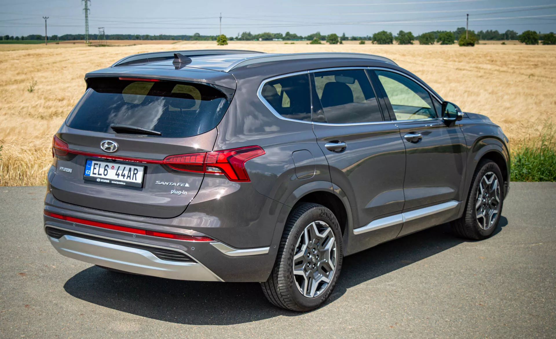 Obrázok zobrazuje trojštvrťový zadný pohľad na moderné SUV Hyundai Santa Fe 2020, zachytené za jasného denného svetla pod čistou oblohou. Auto je mierne naklonené doprava, čo odhaľuje kľúčové prvky ako zadné dvere s logom značky, názvom modelu a úrovňou výbavy, ostro navrhnuté zadné LED svetlá, zliatinové kolesá s viaclúčovým dizajnom, bočné zrkadlá s integrovanými smerovými svetlami a charakterové línie. Okná sú tónované s chrómovaným rámom a zametajúce línie strechy pridáva športový pôvab. Vozidlo stojí na asfaltovom povrchu s poľom a stromami v pozadí, tmavosivý metalický voz vyzerá nový alebo dobre udržiavaný.