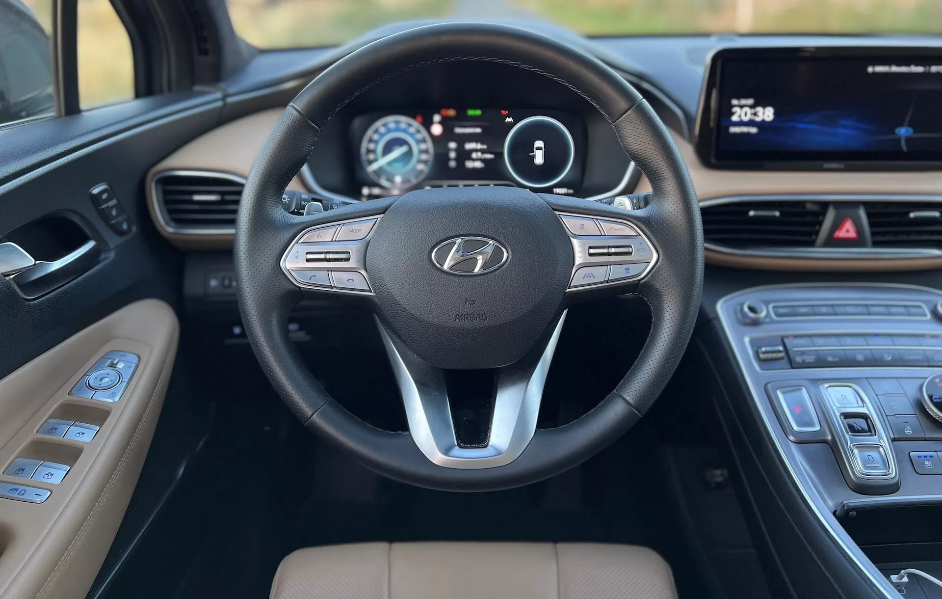 Detailný pohľad zo zadných sedadiel Hyundai Santa-Fe 2020, zameriavajúc sa na predný panel a volant. Trojramenný volant s emblémom Hyundai a ovládačmi pre audio, hlasové príkazy a tempomat. Za volantom je digitálny prístrojový panel zobrazujúci rýchlosť a otáčky na farebnom displeji. Stredová konzola zahrnuje multimediálny dotykový displej a elegantnú klimatizáciu s dotykovými ovládačmi. Radenie má moderný dizajn so striebornými akcentami a svetlohnedé čalúnenie kontrastuje s tmavým panelom a volantom, čo poukazuje na technologicky vyspelý a pohodlný interiér.