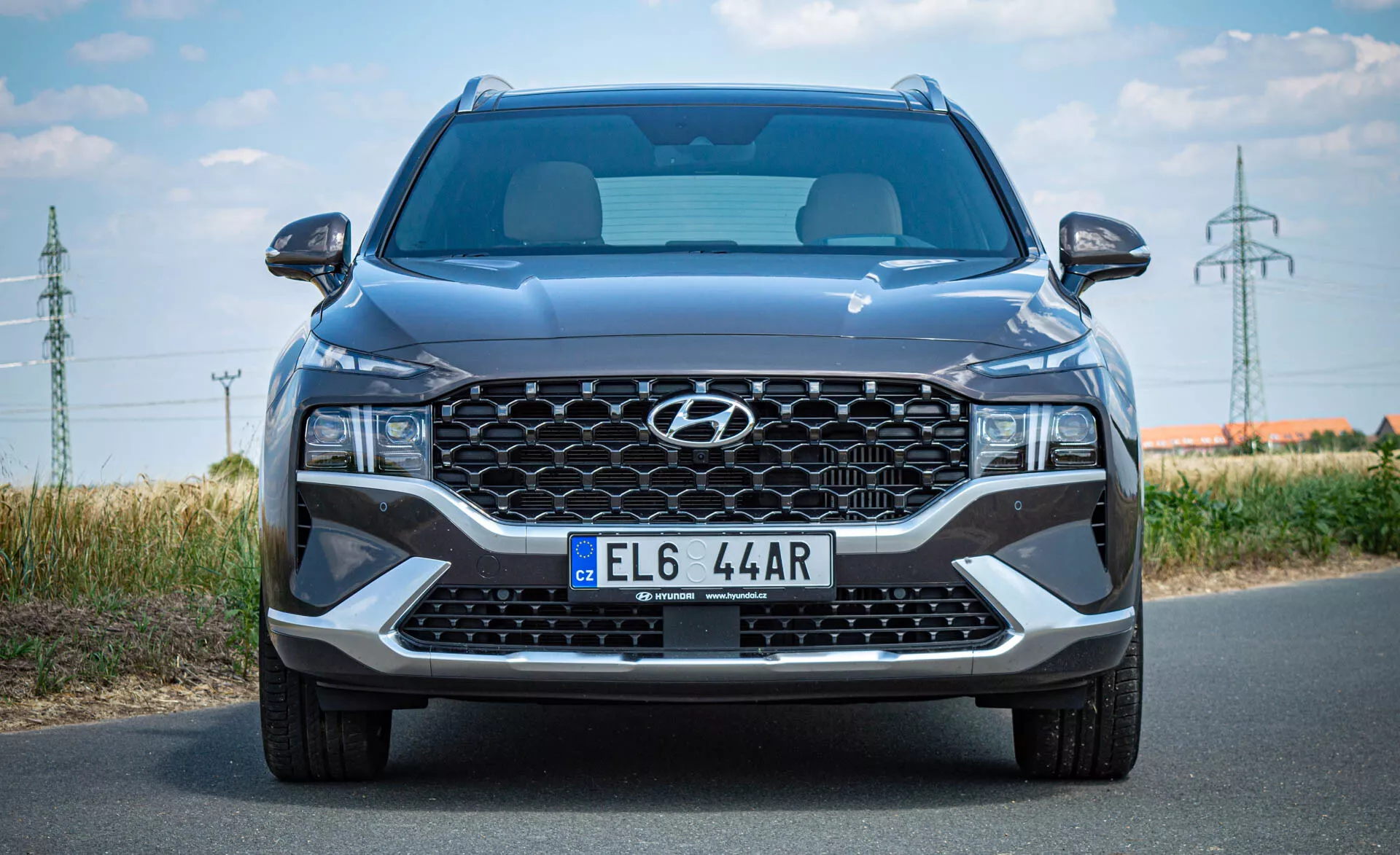 Čelný pohľad na moderné SUV Hyundai Santa Fe 2020 urobený počas denného svetla. Obraz ukazuje veľkú, kaskádovú mriežku s centrálnym logom Hyundai, európsku poznávaciu značku, elegantné uhlové LED svetlomety, široký centrálny prívod vzduchu a strieborné akcenty na dolnom nárazníku. Viditeľné sú tiež tvarované línie kapoty, čiastočné čelné sklo a bočné zrkadlá s integrovanými smerovkami. Obrázok zdôrazňuje veľkosť auta, popisuje jeho moderný dizajn a technologické prvky.