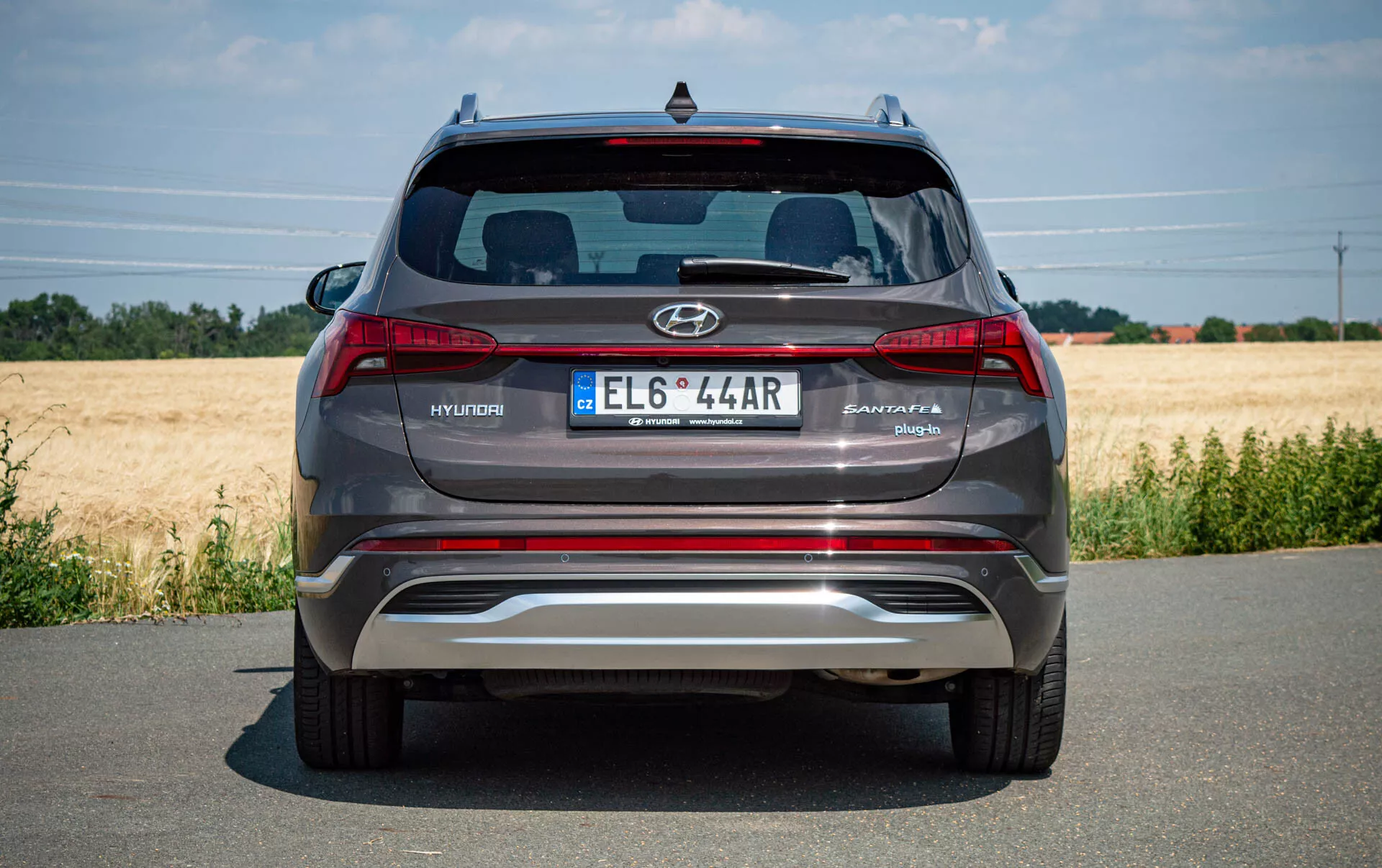 Zadný pohľad na moderné strieborné-šedé SUV Hyundai Santa Fe 2020 zaparkované na dláždenej ceste, zobrazuje logo Hyundai, názov modelu Santa Fe, európsku poznávaciu značku, zadné svetlá, reflexný kovový zadný nárazník s integrovanými parkovacími senzormi, dvojité výfukové špičky, zadné okno so spojlerom Hyundai a tretie brzdové svetlo. Jasné počasie s poľom v pozadí.