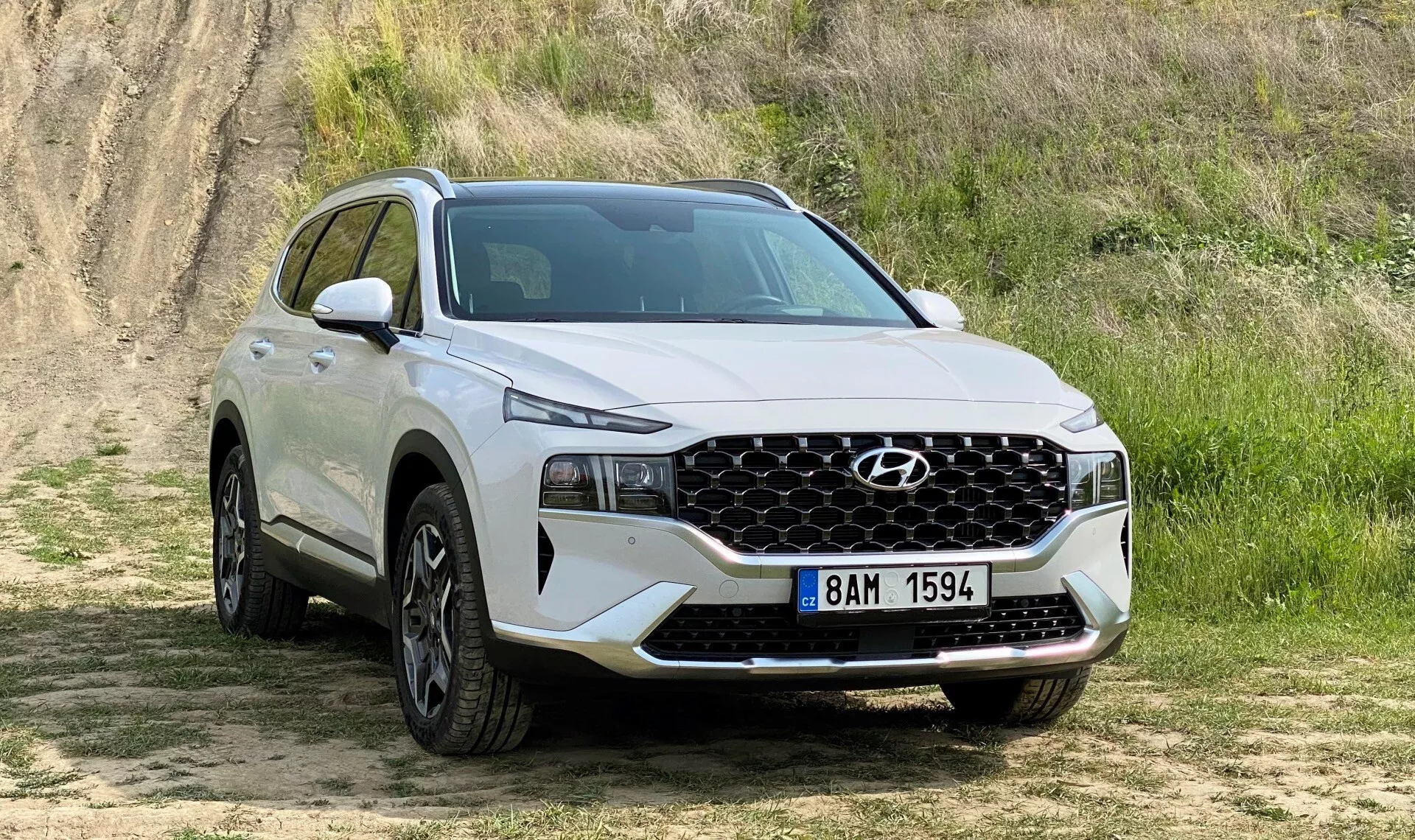 Obrázok zobrazuje biele SUV Hyundai Santa Fe 2020 zaparkované na nezpevnenom povrchu s krátkou trávou a zvýšeným pozadím z hliny, čo naznačuje mierne terénne prostredie. Zachytené z trojštvrtinového predného pohľadu, obrázok jasne ukazuje prednú fasádu vozidla vrátane výraznej geometrickej mriežky, ktorá sa rozkladá takmer cez celú šírku, v strede s logom Hyundai a lemovanú elegantnými LED svetlami. Dolný predný nárazník obsahuje ďalšie osvetlenie alebo senzory. Z bočného profilu až po zadné dvere pasažiera veľké hliníkové kolesá vyplňujú oblúky kolies, a nad nimi je práca, ktorá naznačuje robustnosť. Silueta SUV ukazuje klesajúcu líniu strechy, ktorá je typická pre moderný športový dizajn. Scéna je prirodzene osvetlená a zvýrazňuje vlastnosti SUV bez ostrých tieňov.