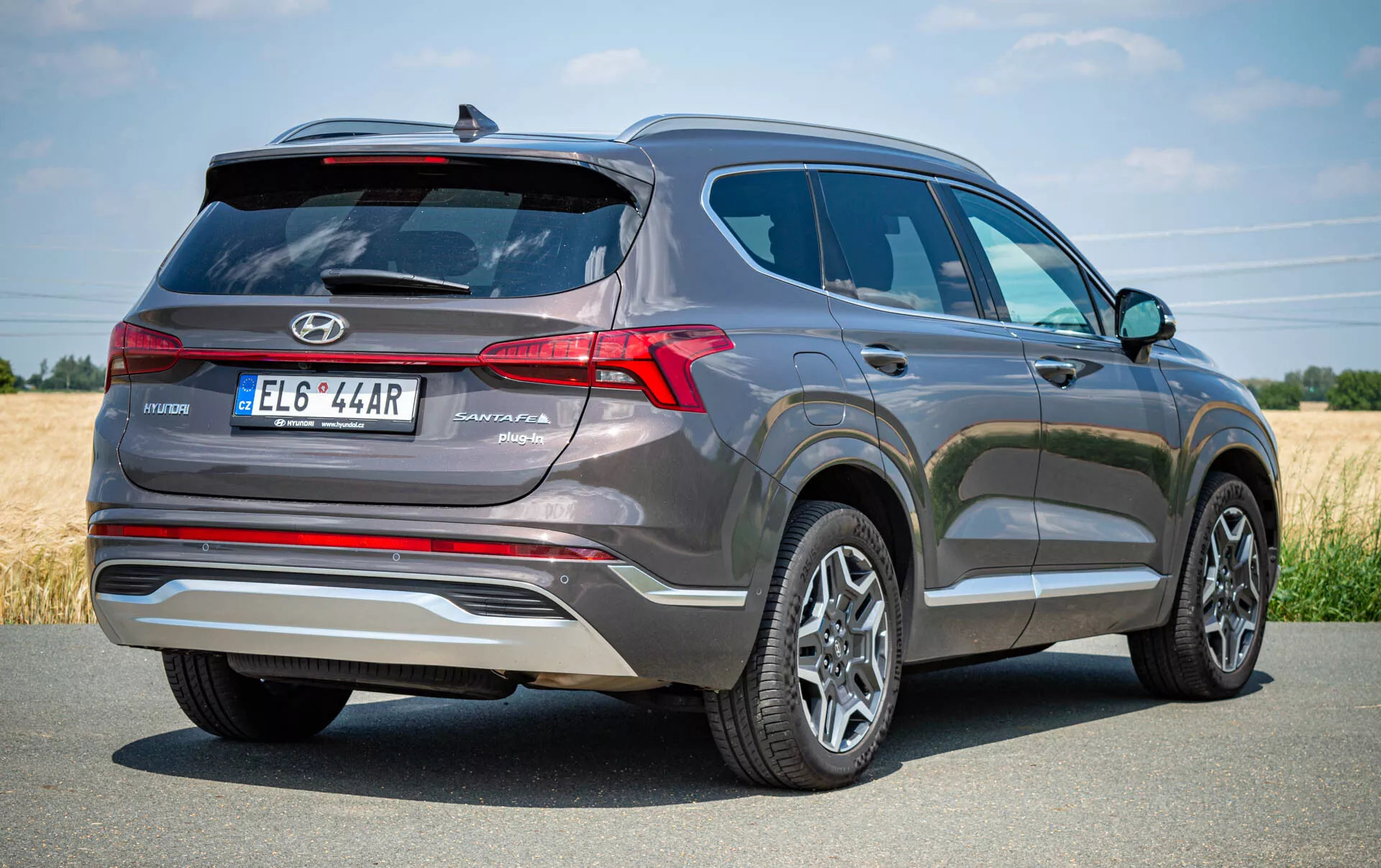 Trojštvrtinový pohľad zozadu na sivé SUV Hyundai Santa Fe 2020, zdôrazňujúci zadnú ľavú stranu vozidla. Obrázok ukazuje výrazné LED zadné svetlá, zadný ostrekovač, odmrazovač a stierač, s emblémom Hyundai a názvom modelu 'Santa Fe' pri stredovo umiestnenej tabuľke s evidenčným číslom. Strieborný lem pridáva štýl a nad evidenčným číslom sa nachádza parkovacia kamera alebo senzorový systém. Viditeľné sú aj viaclúčové zliatinové kolesá a pneumatiky v dobrom stave, čo naznačuje moderné, dobre udržiavané vozidlo. Ideálne na automobilové inzeráty, recenzie alebo obsah zameraný na technológiu.