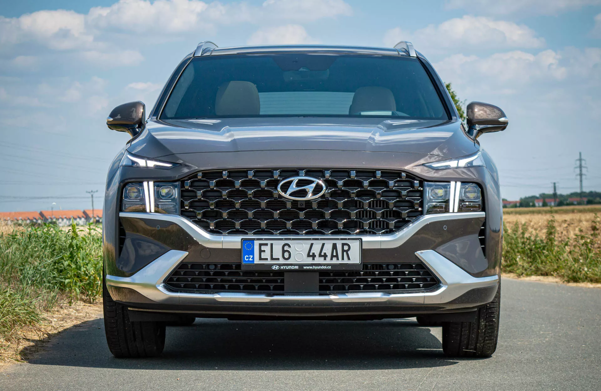 Predný pohľad na SUV Hyundai Santa-Fe 2020 na asfaltovej ceste, ukazujúci veľkú šesťuholníkovú mriežku so vzorom včelieho plástu, elegantné LED svetlomety, centrálne umiestnený znak na hornej časti mriežky, LED denné svietenie pod hlavnými svetlometmi a európsku štýlovú poznávaciu značku na spodnej časti mriežky. Pozadie tvorí rozmazaná vidiecka alebo polovidiecka krajina s jasnou oblohou, čo naznačuje dobré počasie.