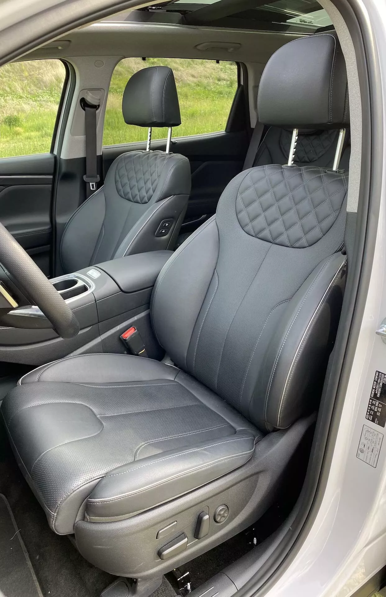 Interiér Hyundai Santa Fe 2020 ukazujúci sedadlo vodiča z otvorených dverí. Blízky vertikálny záber zachytáva predné sedadlá, zameriavajúc sa na sedadlo vodiča s tmavým koženým čalúnením so vzorom prešívaných diamantov na operadle a sedadle, čo naznačuje prémiovú verziu. Viditeľné elektronické ovládače naznačujú nastaviteľné polohy sedadla. Opierku hlavy možno upraviť a panel dverí má ovládače okien a pravdepodobne aj zrkadiel. Interiér obsahuje detaily s kontrastným prešívaním, čím sa zvyšuje pocit luxusu.