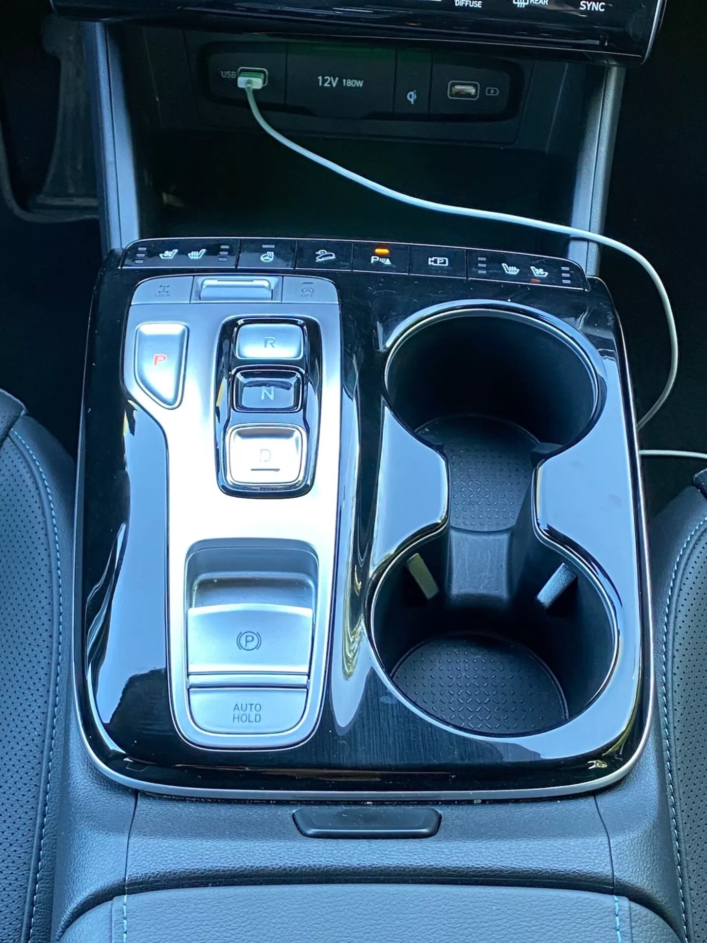 Obrázok zobrazuje centrálnu konzolu Hyundai Tucson 2020 s elektronickým voličom prevodovky v polohe 'N' obklopený lesklým čiernym dekorom. Naľavo sú ovládače vrátane tlačidla napájania, voličov jazdných režimov a funkcie 'AUTO HOLD'. Napravo sú dve držiaky na poháre, pričom v jednom je biely nabíjací kábel. V hornej časti je vidieť časť informačného displeja s indikátormi USB portov, čo poukazuje na moderné technológie a luxus vozidla.