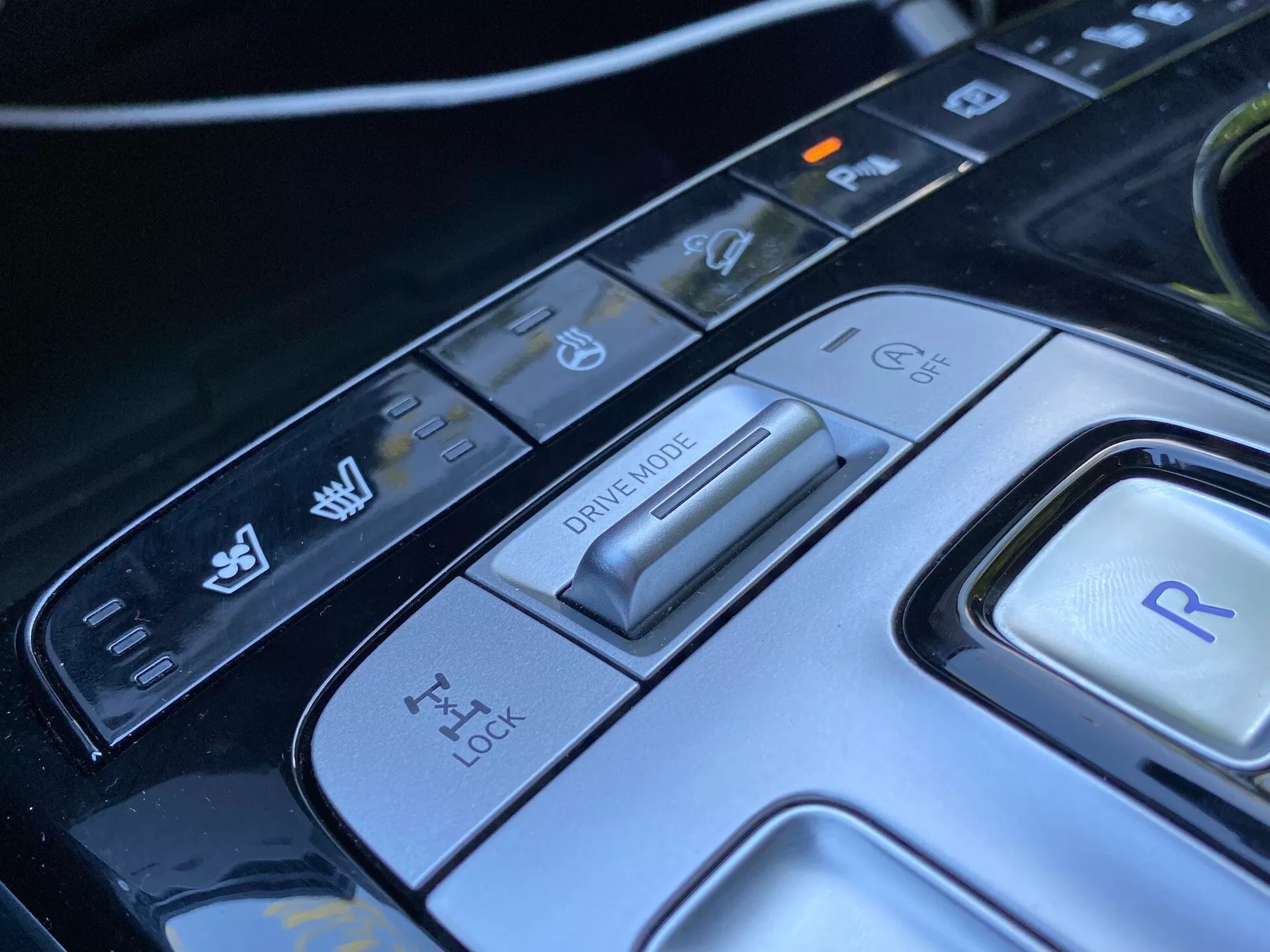 Detailný pohľad na stredovú konzolu vozidla Hyundai Tucson 2020, zameraný na volič jazdného režimu a rôzne ovládacie tlačidlá. Obraz ukazuje pokročilý prepínač jazdného režimu pre rôzne jazdné podmienky, panel pre kontrolu trakcie, tlačidlo pre spiatočku a ďalšie ovládacie prvky pre stabilitu vozidla a asistenčné funkcie parkovania. Elegantný dizajn a high-tech materiály zdôrazňujú sofistikovaný automobilový interiér tohto luxusného moderného auta.