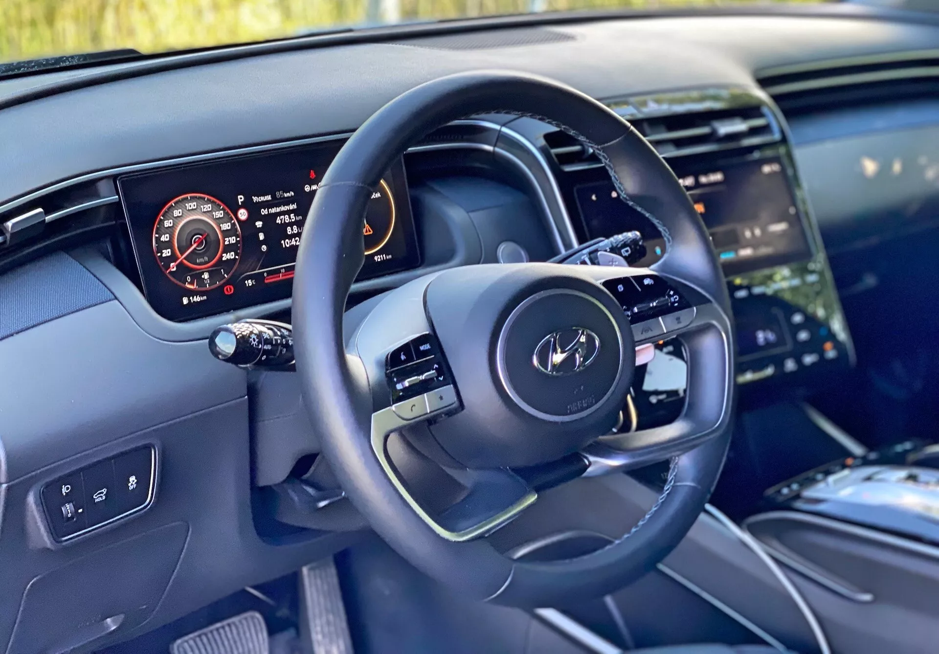 Pohľad na interiér Hyundai Tucson 2020 z perspektívy vodiča, ktorý ukazuje moderný volant s multifunkčnými ovládačmi a digitálny prístrojový panel zobrazujúci rýchlomer, otáčkomer, hladinu paliva a teplotu motora. Obrázok tiež zdôrazňuje časť stredovej konzoly s veľkým dotykovým infotainment systémom, čistý dizajn palubnej dosky s ambientným osvetlením a vysoko kvalitné materiály naznačujúce luxusné povrchy.
