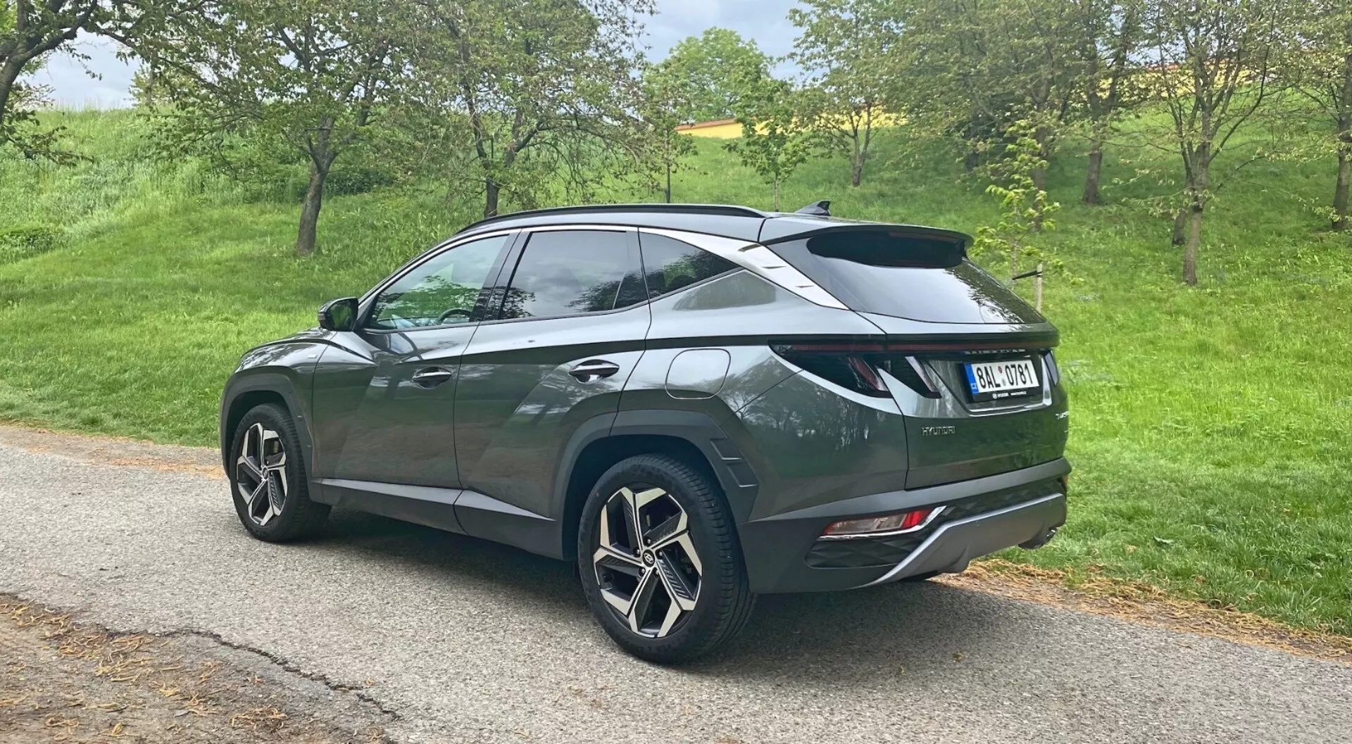 Pohľad na tri štvrtiny zozadu na Hyundai Tucson 1.6 T-GDi Pure SUV z roku 2020 zaparkovaný na úzkej ceste v vidieckom prostredí, ktorý ukazuje veľké viacramenné zliatinové kolesá, tmavý metalický lak s klesajúcou líniou strechy, zadný spojler, zatmavené okná vzadu, moderné zadné svetlá a dvojité výfukové koncovky integrované do zadného nárazníka.
