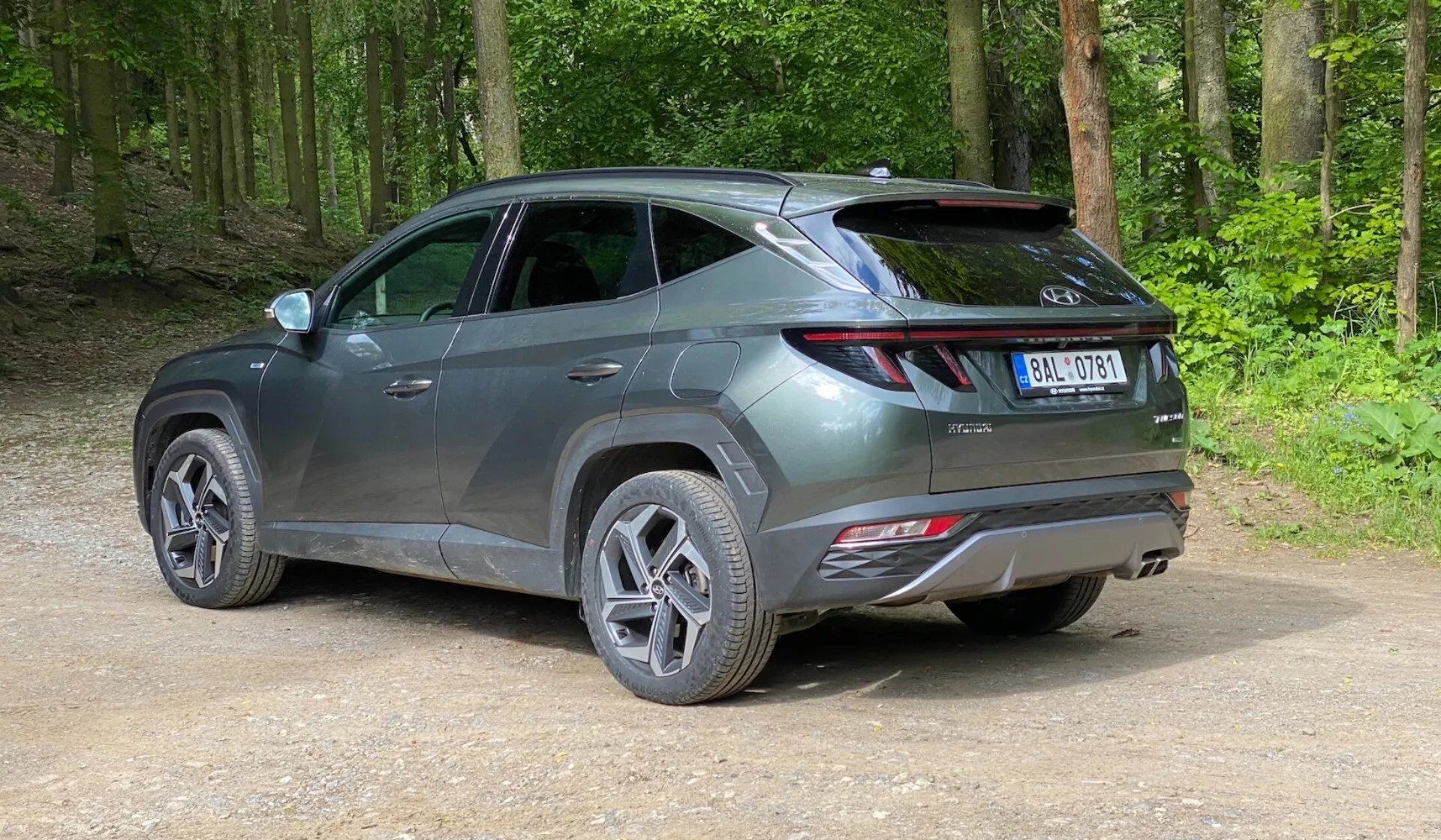 Moderné SUV zaparkované na nespevnejej ceste v lesnej oblasti, zachytené z trojštvrtinového zadného pohľadu z mierneho zvýšenia. Vozidlo má elegantný, súčasný dizajn s ostrými líniami, predĺženými zadnými svetlami obopínajúcimi zozadu po boky, viditeľným spojlerom a reflektormi na zadnom nárazníku, čo naznačuje pokročilé bezpečnostné funkcie. Vozidlo v metalickej šedej farbe má viaclúčové zliatinové kolesá a cestné pneumatiky, s lesklým povrchom naznačujúcim, že je nové alebo dobre udržiavané. Okolo auta je zelený, bujný les so zhutnenou hlinou a voľným štrkom na zemi.