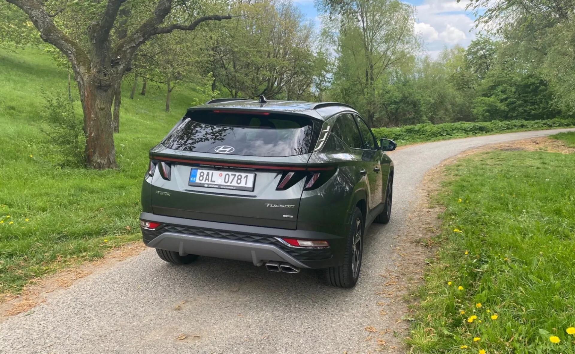 Pohľad na zadnú časť moderného SUV Hyundai Tucson 2020 zaparkovaného na úzkej asfaltovej ceste s prírodným trávnatým pozadím, čo naznačuje vidiecke alebo parkové prostredie. Obrázok zachytáva ľavú zadnú stranu z mierne nízkeho uhla, zameriava sa na dizajnové prvky, ako je tvarovaný zadný nárazník, dvojité výfukové koncovky, charakteristické LED zadné svetlá a integrovaný strešný spojler. Logo Hyundai a názov modelu 'Tucson' sú jasne viditeľné pod zadným sklom. Fotografia ukazuje dobre udržiavané vozidlo s detailmi ako vysoko umiestnené brzdové svetlo a čiastočne viditeľné moderné hliníkové kolesá. Pozadie obsahuje jemne rozmazané stromy a lístie pod zamračenou oblohou, ktorá poskytuje rozptýlené prirodzené svetlo.