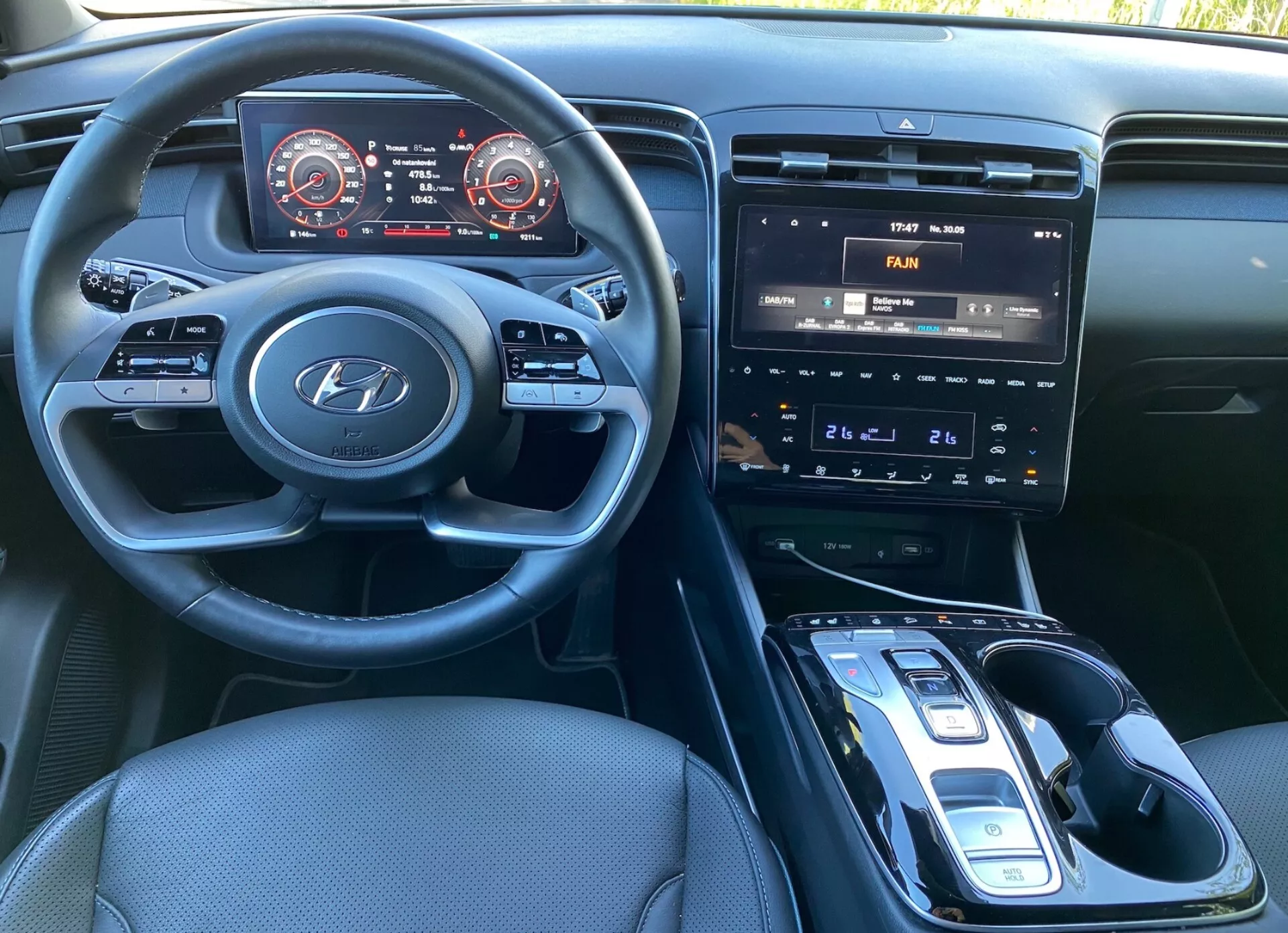 Pohľad na interiér Hyundai Tucson 2020 zo sedadla vodiča, ukazujúci volant s logom Hyundai a ovládacími prvkami pre zvukový systém, telefon a tempomat. Prístrojová doska obsahuje digitálny panel s tachometrom a rýchlomerom s červeným zvýraznením. Stredová konzola má infotainment systém s dotykovou obrazovkou zobrazujúci ovládanie klimatizácie, fyzické tlačidlá a gombíky na ovládanie klimatizácie a automatickú radiacu páku s elektronickým voličom. Je vidieť potenciálnu bezdrôtovú nabíjačku, tmavé textilné čalúnenie a dobre osvetlený interiér.