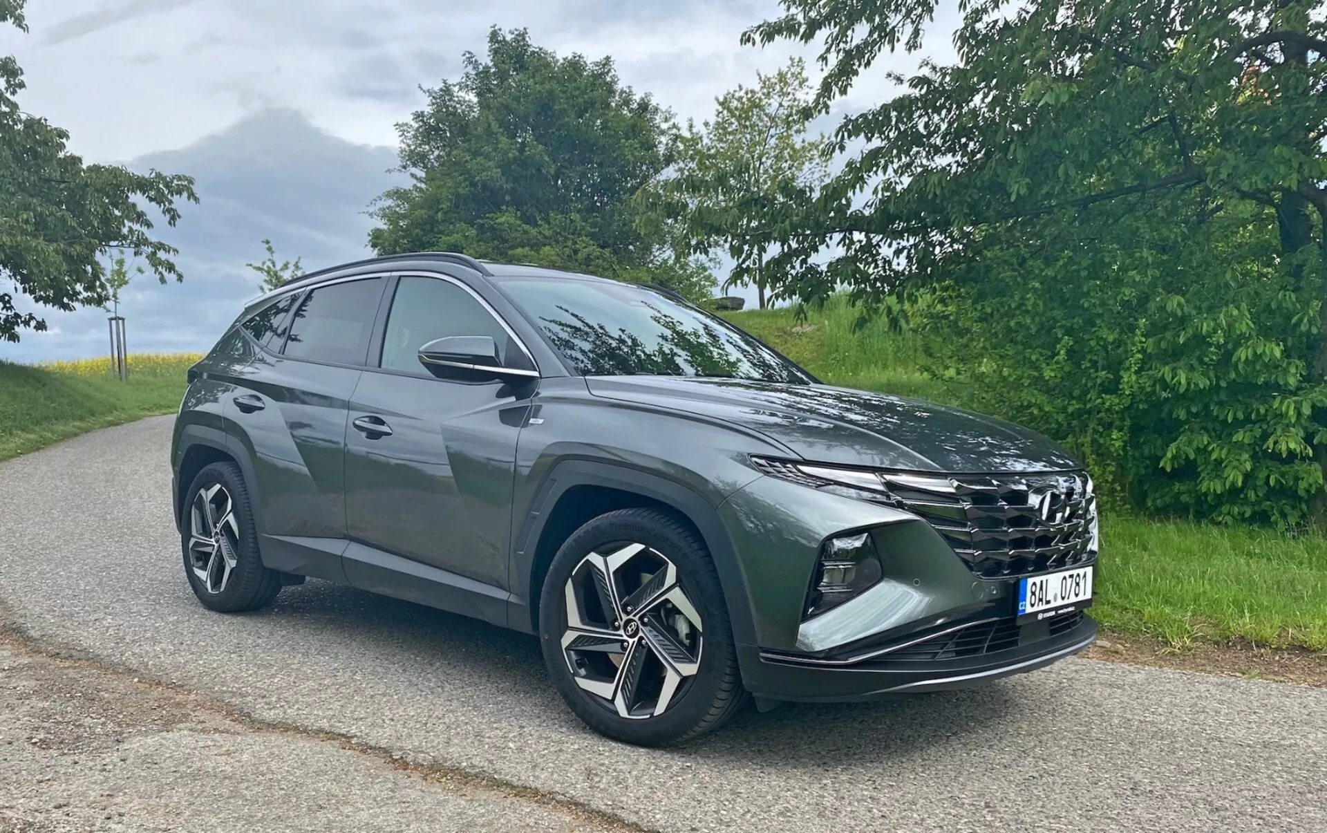 Obrázok zobrazuje moderné SUV Hyundai Tucson 1.6 T-GDi Pure zaparkované na asfaltovej ceste s prírodným pozadím. Vozidlo je fotografované z predného trojštvrťového uhla, čo zviditeľňuje jeho výraznú mriežku chladiča, elegantné svetlomety, predný nárazník s hmlovými svetlami alebo ventilačnými otvormi, veľké zliatinové kolesá s viaclúčovým dizajnom a spätné zrkadlá. Silueta SUV sa smerom dozadu sklání, čo naznačuje športový, aerodynamický dizajn. Snímka bola zhotovená v mäkkých denných svetelných podmienkach, čo zvýrazňuje farbu a detaily vozidla bez ostrých tieňov.