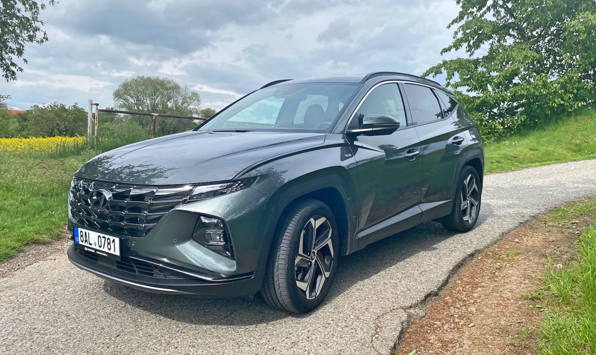 Hyundai Tucson 2020 1.6 T-GDI Pure SUV, zobrazený z predného trojštvrtinového uhla na dláždenej ceste s stromami a trávou v pozadí. Obraz ukazuje masku chladiča vozidla, ostré LED svetlomety, zliatinové kolesá, spätné zrkadlá s integrovanými smerovkami a tmavý metalický lak. Viditeľné sú predné vodičove a zadné dvere. Fotografia bola zhotovená počas dňa za zatiahnutých svetelných podmienok.