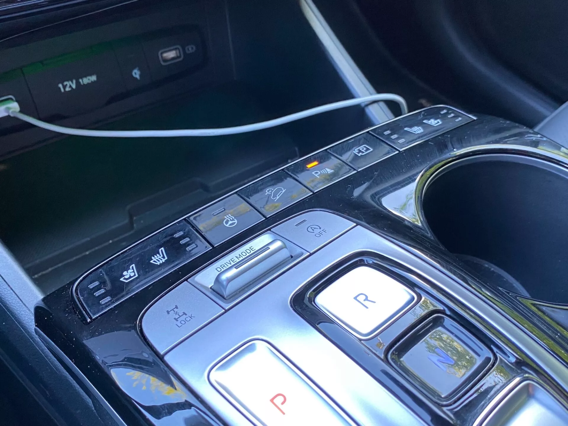 Detailný pohľad na stredovú konzolu a oblasť výberu prevodov vozidla Hyundai Tucson 2020 1.6 T-GDi Pure, so moderným dizajnom s tlačidlami na parkovanie, spätný chod, neutrál, jazdu a režimy brzdenia/regenerácie, a ďalšími tlačidlami na deaktiváciu funkcie auto start/stop, parkovacie senzory/automatické parkovanie a varovanie pred opustením jazdného pruhu. Viditeľný multimediálny systém, držiaky na nápoje a 12V zásuvka naznačujú komfort, komfort a pokročilé systémy asistencie vodiča (ADAS). Konzola má elegantný dizajn s lesklým čiernym materiálom a kovovými akcentmi, čo naznačuje luxusný vzhľad.