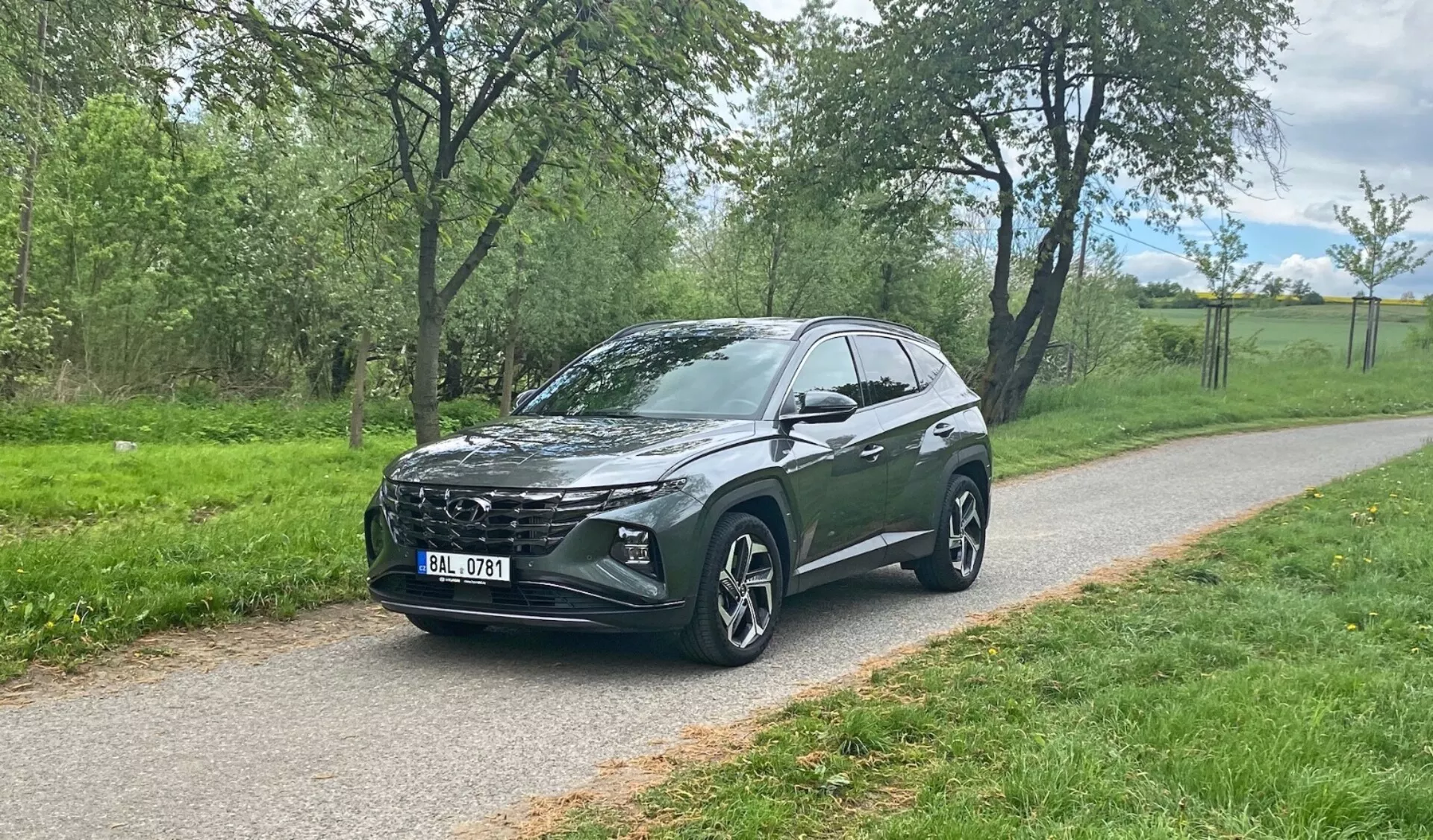 Trojštvrťový predný pohľad na crossover SUV Hyundai Tucson 2020, zaparkovaný na dláždenom chodníku so zeleňou v pozadí, ukazujúci výraznú prednú mriežku vozidla, elegantné LED svetlomety, veľké zliatinové kolesá s viac lúčmi, sklonenú strechu dozadu, vysokú svetlú výšku, integrované bočné zrkadlá, tónované sklá a štýlové línie karosérie.