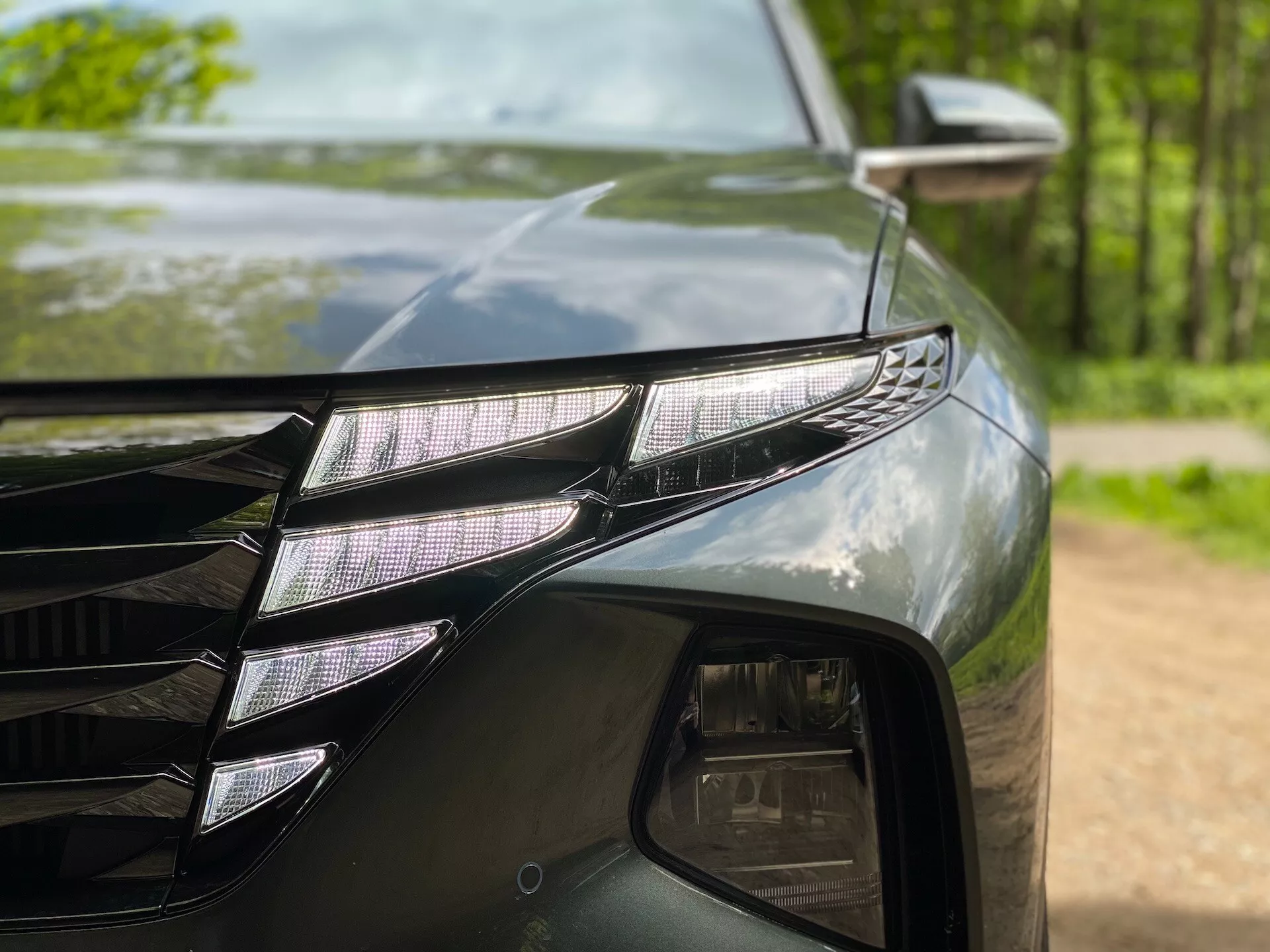 Detailný záber ľavej prednej časti Hyundai Tucson 2020 (Hyundai-Tucson-1-6-T-GDi-Pure), predstavuje LED svetlomet s jednotlivými LED projektormi a výrazným LED DRL prúžkom. Tmavý, lesklý lak odráža okolité stromy. Viditeľná je tiež časť mriežky s hranatými čiarami a bočné zrkadlo s indikátorom smeru.