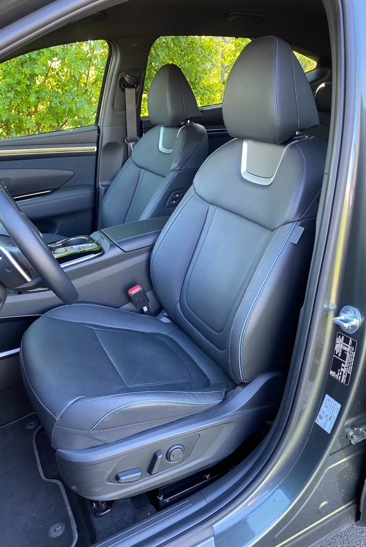 Interiér Hyundai Tucson 2020 1.6 T-GDI Pure zobrazujúci predné sedadlo spolujazdca s čiernym koženým čalúnením, detailným prešitím a ovládacími prvkami elektrického nastavenia. Viditeľná stredová konzola s držiakom na poháre, dverový panel so zhodnými materiálmi, madlo, úložný priestor a mriežka reproduktora naznačujúca pokročilý audiosystém. Čiastočne viditeľná palubná doska a priehradka na rukavice. Koberec podobný podlahovej rohoži a mierny odraz slnečného svetla na dverovom paneli.
