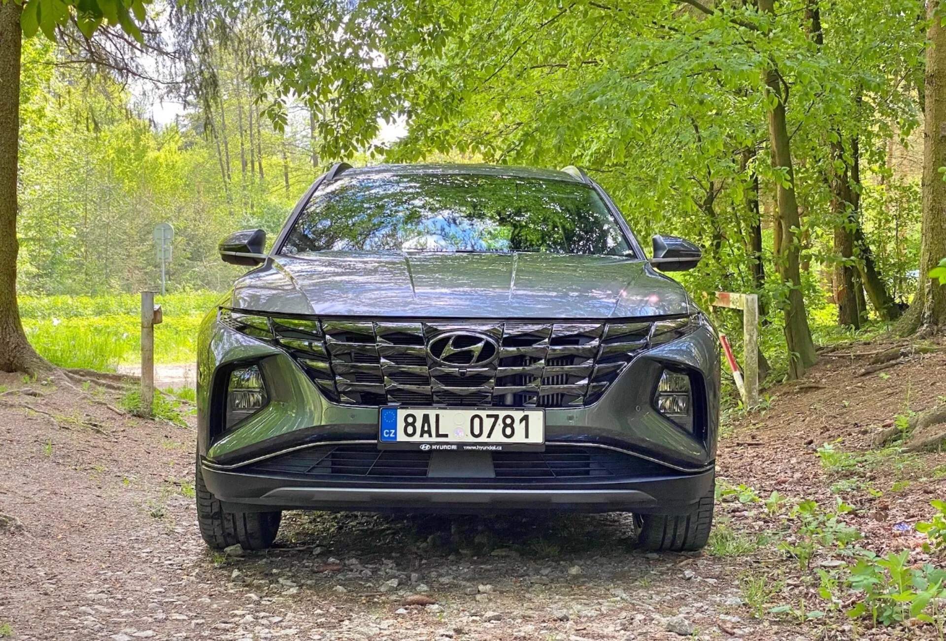 Predný pohľad na vozidlo Hyundai Tucson 1.6 T-GDi Pure 2020, zobrazujúci nízkoúhlý záber zameraný na mriežku, predný nárazník a oblasť kapoty. Auto je zaparkované na štrkovom chodníku v prírodnom prostredí, čo naznačuje jeho vhodnosť pre vonkajšie prostredia. Kľúčové vlastnosti zahŕňajú výrazný dizajn mriežky, elegantné LED svetlomety a viditeľné logo Hyundai.