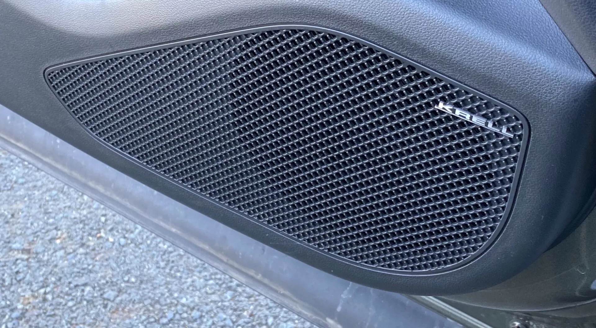 Detailný bočný pohľad na Hyundai Tucson 2020 1.6 T-GDI Pure, zameraný na bočnú mriežku so vzorom sieťky s diamantovými výrezmi. Mriežka, zapustená do tvarovaného obrysu, je zakomponovaná do panelu karosérie auta s matným povrchom, obklopená hladkým zakriveným povrchom. Obrázok, fotený z uhlu zdola nahor, ukazuje prirodzené vonkajšie osvetlenie s kúskami asfaltu a cestnými nečistotami, ktoré podčiarkujú elegantný, výkonný vzhľad.
