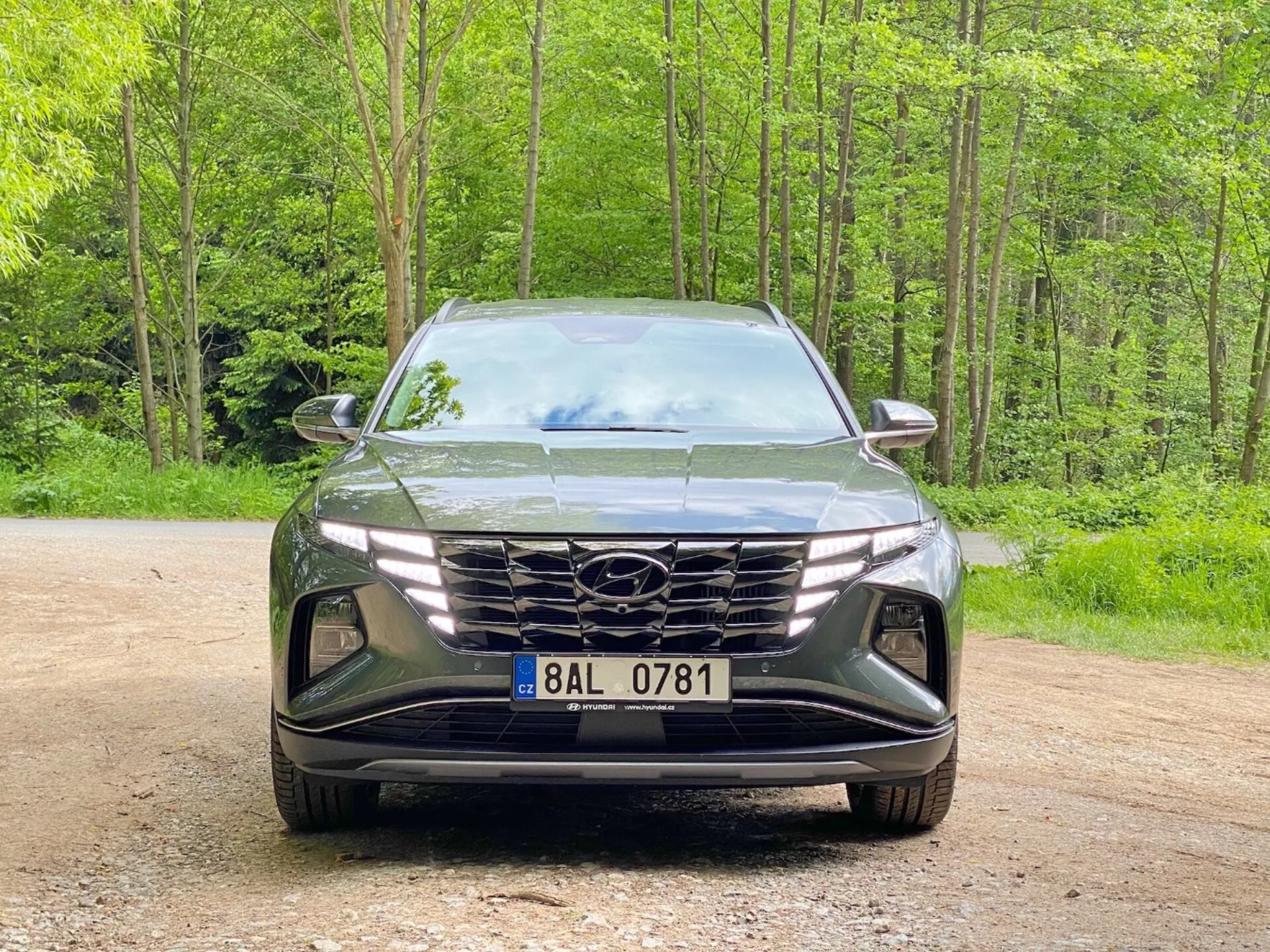 Čelný pohľad na Hyundai Tucson 2020 (Hyundai Tucson 1.6 T-GDI Pure) moderné SUV zaparkované na štrkovej ceste s lesnatým pozadím. Obrázok zachytáva elegantný, agresívny dizajn s veľkou geometrickou mriežkou s logom výrobcu, úzkymi uhlovými LED svetlometmi a tvarovaným predným nárazníkom s nasávacími otvormi a priestorom pre hmlové svetlá, zdôrazňujúci pokročilú aerodynamiku a osvetľovaciu technológiu.