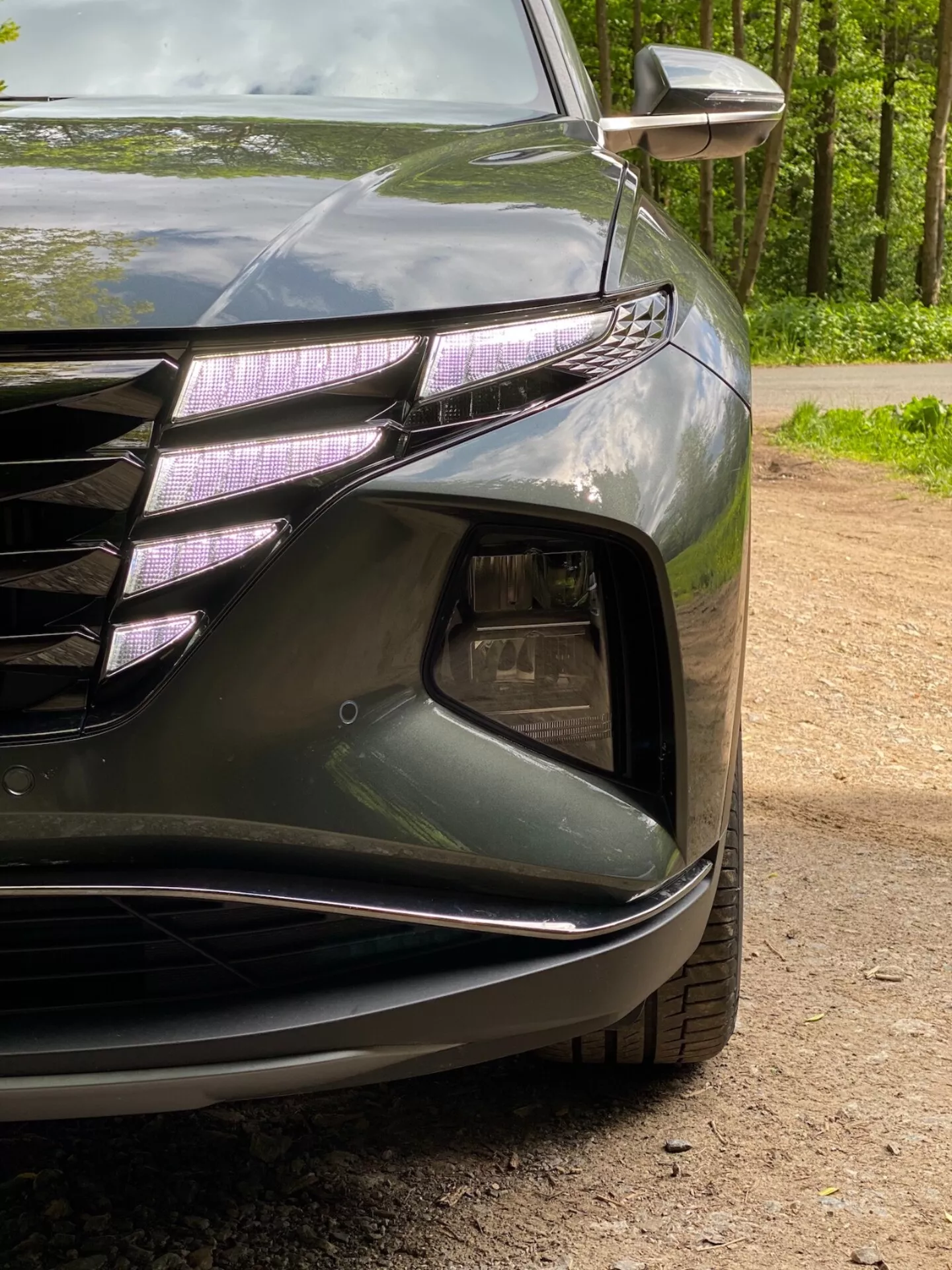 Zblízka pohľad na predný ľavý roh Hyundai Tucson 2020 1.6 T-GDI Pure, zobrazujúci svetlomet s LED denným svietením a projektorovým objektívom, časť prednej mriežky s horizontálnymi čiarami a chrómovými akcentmi, sekciu prívodu vzduchu s krytom hmlových svetiel, tvarovaný predný nárazník s hladkými líniami a integrované smerové svetlo v zrkadle. Tmavý lesklý lak zvýrazňuje aerodynamické krivky vozidla.
