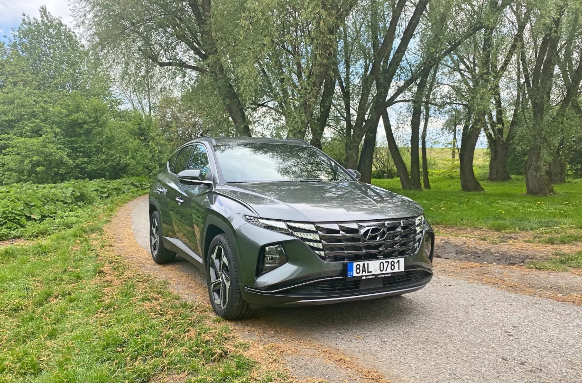 Trojštvrťový pohľad zpredu na Hyundai Tucson 1.6 T-GDi Pure 2020, ktorý ukazuje moderné SUV prvky ako elegantné LED svetlomety, výrazná geometrická predná mriežka, aerodynamická kapota, zliatinové kolesá, bočné zrkadlá s integrovanými smerovými svetlami a metalická farba, na pozadí úzkej asfaltovej cesty so stromami, čo zdôrazňuje jeho všestrannosť pre mestské aj vidiecke prostredie.