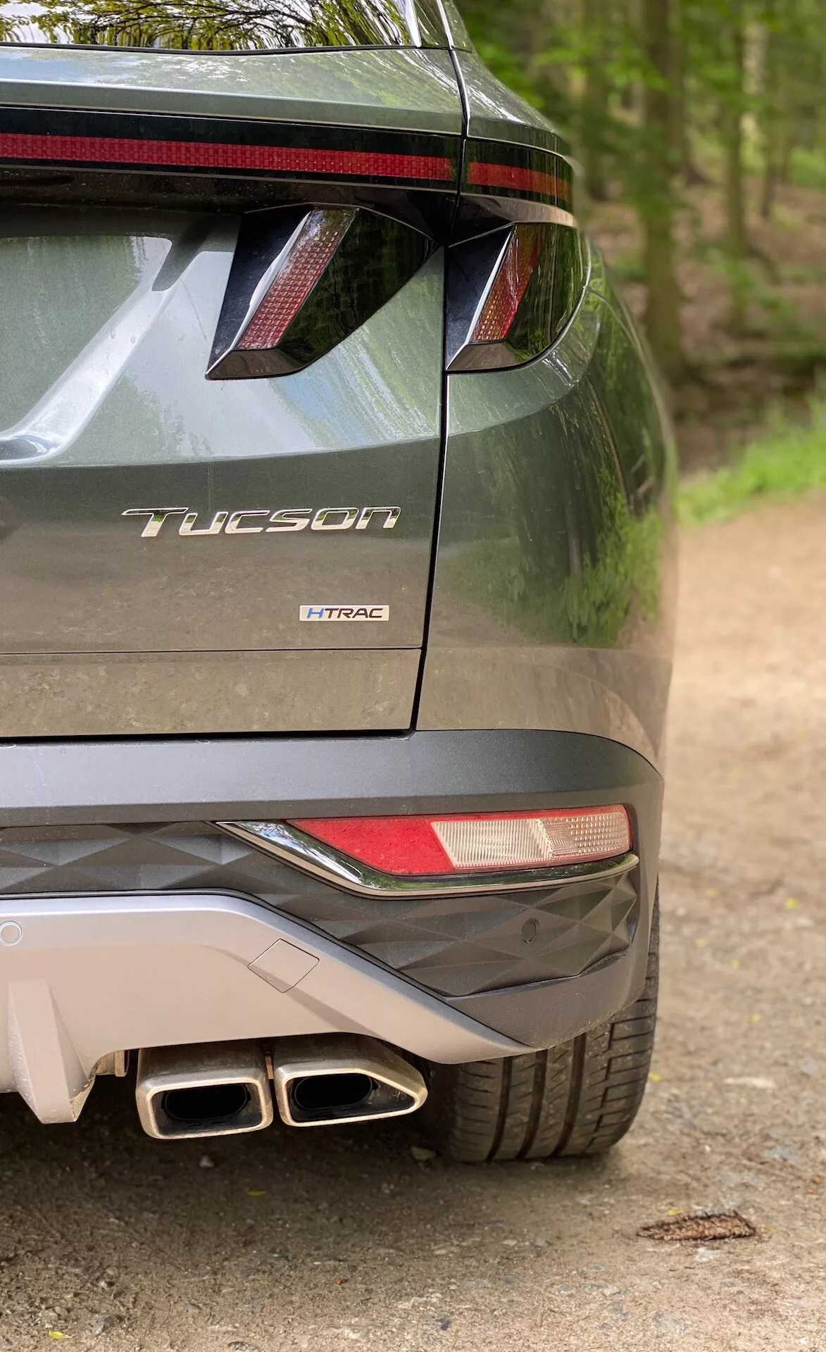 Čiastočný zadný pohľad na model Hyundai Tucson 2020, ktorý naznačuje, že ide o verziu 1.6 T-GDi Pure, zobrazujúci pravú stranu nárazníka, čiastočné zadné svetlo, zadný spojler s LED brzdovým svetlom, dvojité výfukové koncovky a označenie modelu s názvom 'Tucson' a technológiu motora 'T-GDi'.