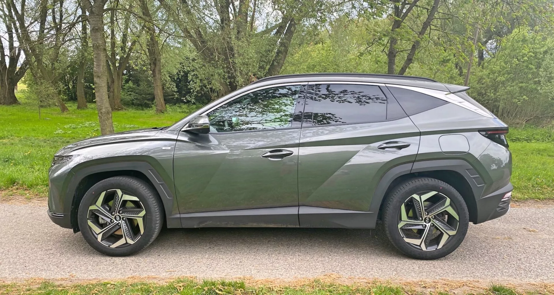 Bočný profil moderného SUV Hyundai Tucson 1.6 T-GDI Pure 2020 zaparkovaného na dláždenej ceste vedľa trávnatej plochy s stromami. Predstavuje elegantný, aerodynamický dizajn so sklonenou strešnou líniou, výraznou líniou karosérie, veľkými viacpaprskovými zliatinovými kolesami s nízkoprofilovými pneumatikami, tónovanými okienkami s viditeľným B-stĺpikom, zapustenými rukoväťami dverí, farebnými bočnými zrkadlami a chrómovým lemovaním pozdĺž okienka. Vozidlo zachytáva moderný štýl a potenciálne pokročilé funkcie z bočného uhla.